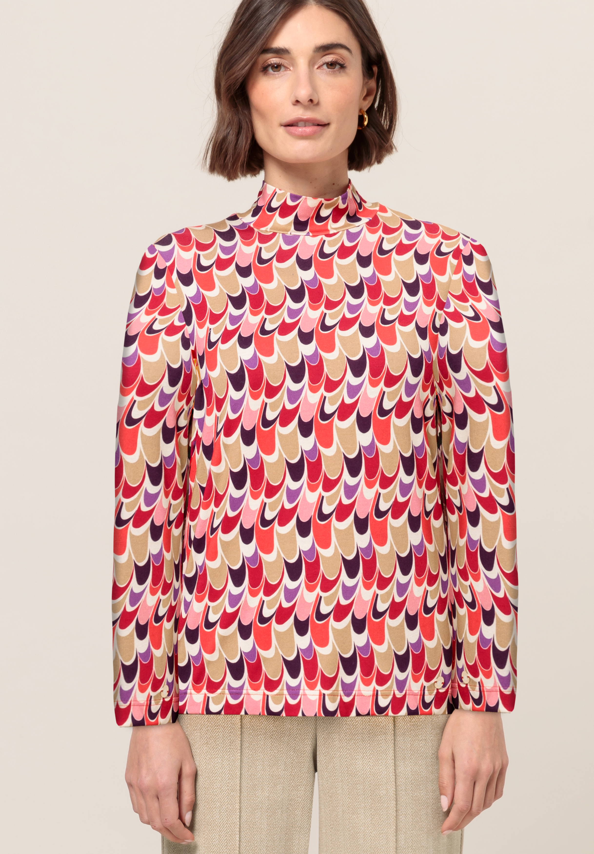 Frau trägt eine langärmelige Bluse mit einem farbenfrohen, wellenförmigen geometrischen Muster in Rot-, Rosa-, Beige- und Lilatönen. Sie steht nach vorne gerichtet, mit neutralem Gesichtsausdruck und hellem Hintergrund.