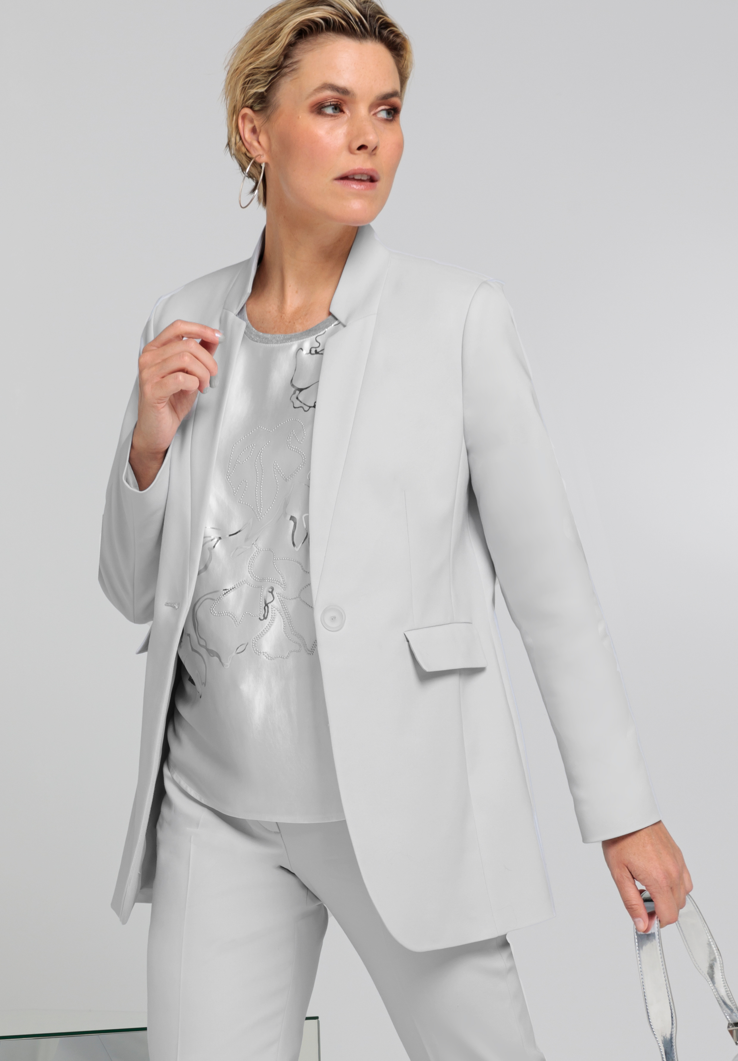 Eine Frau mit kurzen blonden Haaren trägt einen hellgrauen Anzug mit passendem Blazer und Hose über einer glänzenden silbernen Bluse und steht vor einem schlichten grauen Hintergrund.