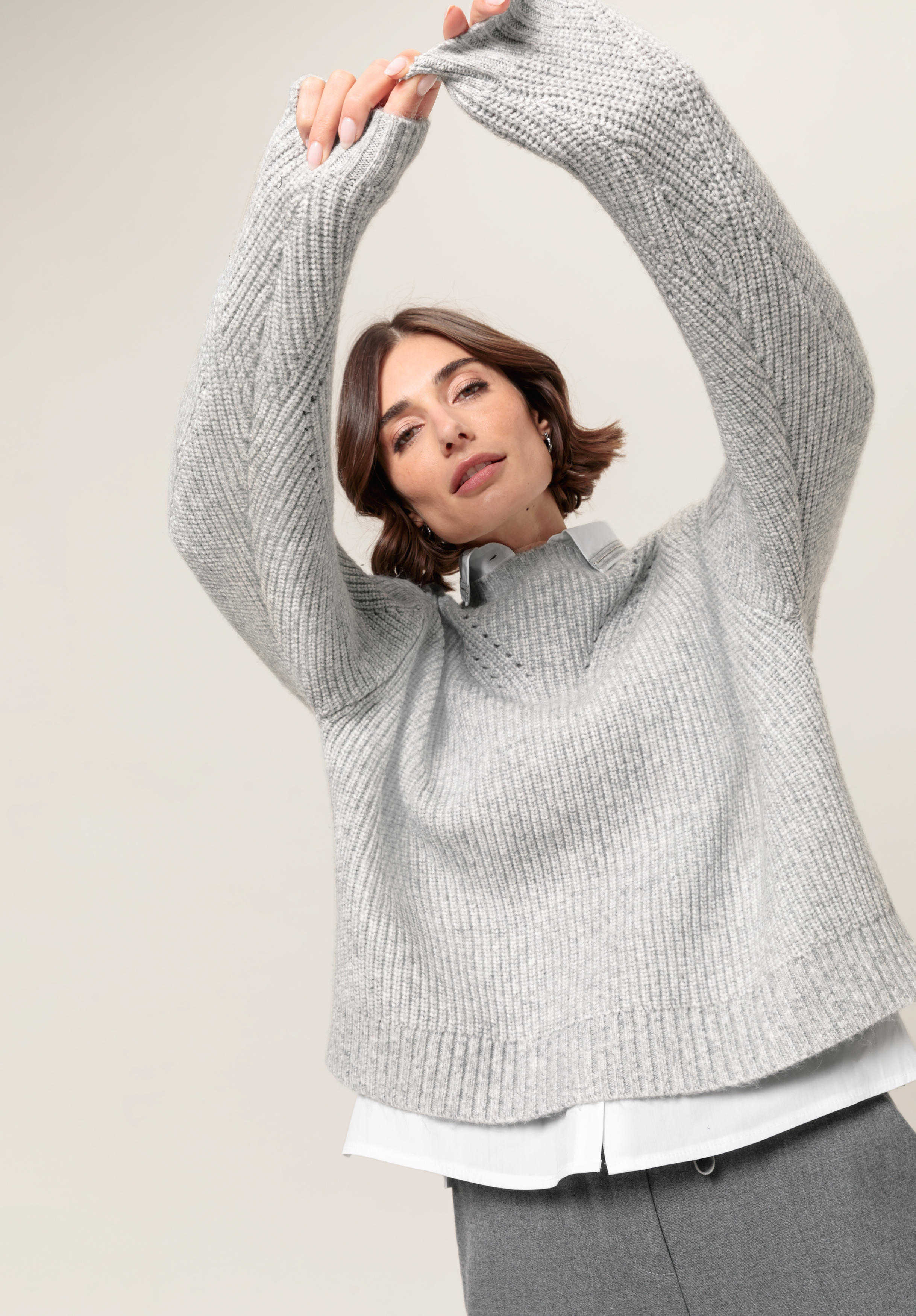 Eine Frau in einem hellgrauen, übergroßen Strickpullover und einer grauen Hose posiert mit erhobenen Armen vor einem schlichten Hintergrund. Sie hat kurzes braunes Haar und einen neutralen Gesichtsausdruck.