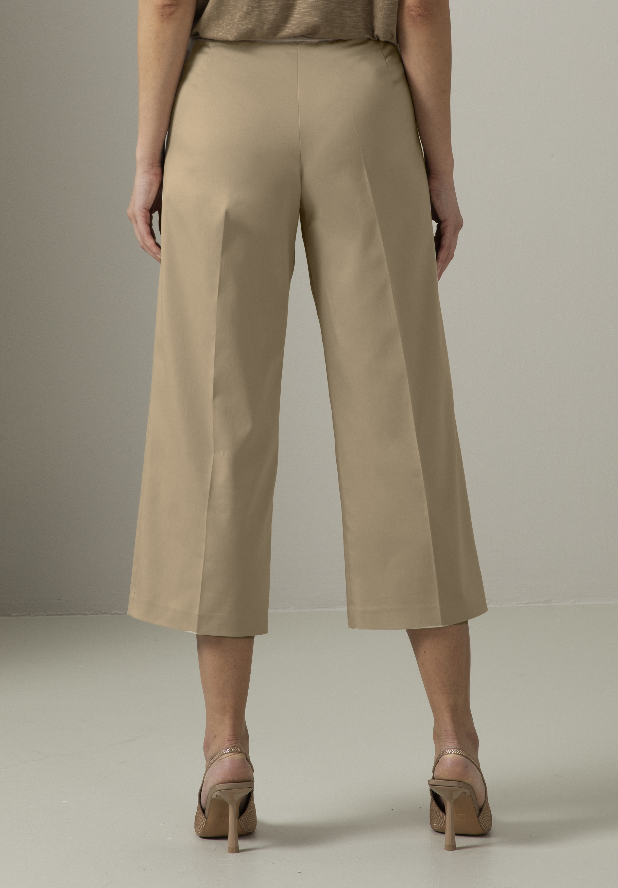 Eine Frau steht mit abgewandtem Gesicht da und trägt eine hellbeige, weite, verkürzte Hose und taupefarbene Sandalen mit Absatz. Die Hose hat klare Linien und endet über den Knöcheln. Der Hintergrund ist schlicht und neutral.