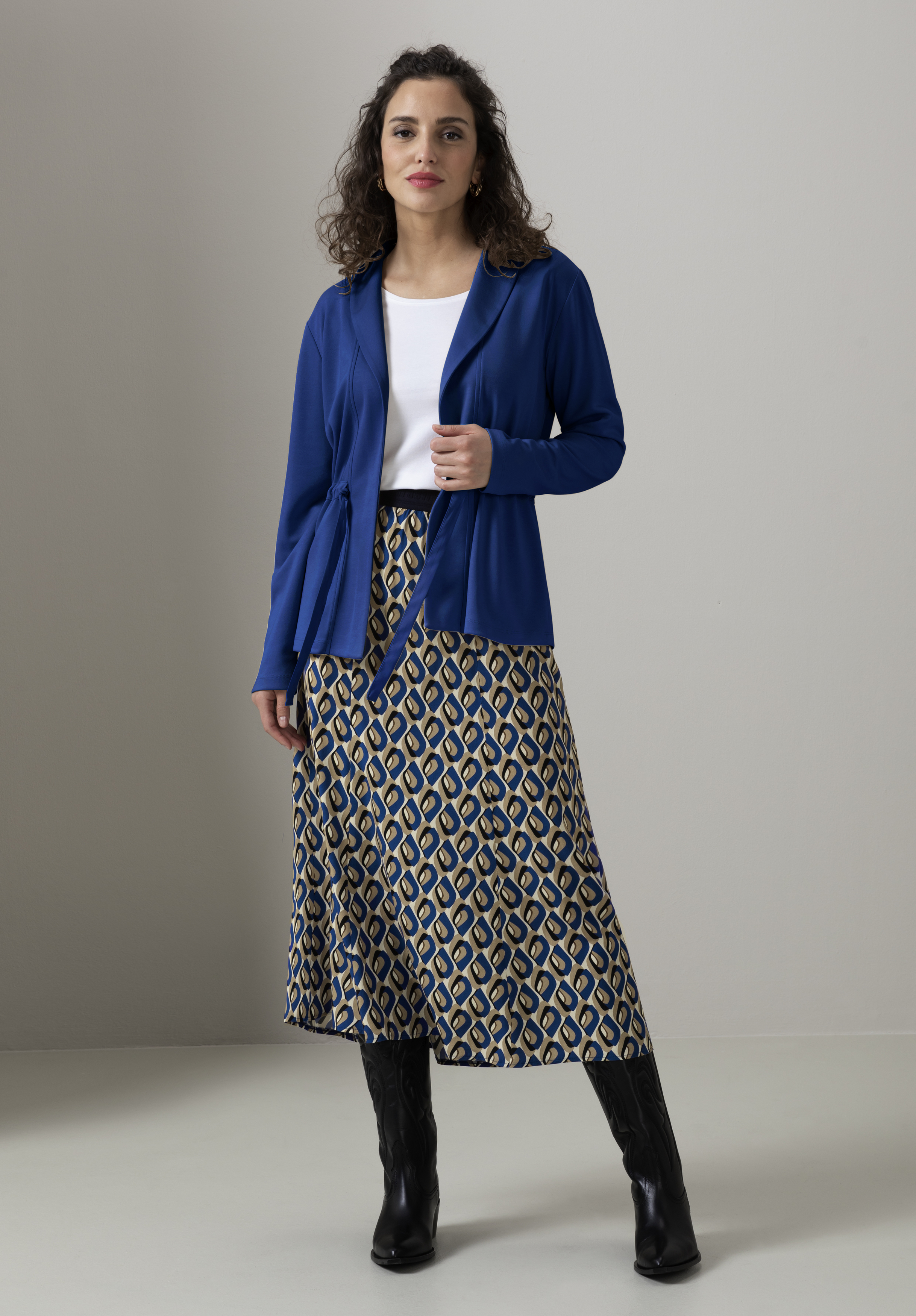 Eine Frau steht vor einem neutralen Hintergrund. Sie trägt eine blaue Jacke über einem weißen Oberteil, einen gemusterten Midirock und schwarze Stiefel. Sie hat lockiges Haar und blickt entspannt in die Kamera.