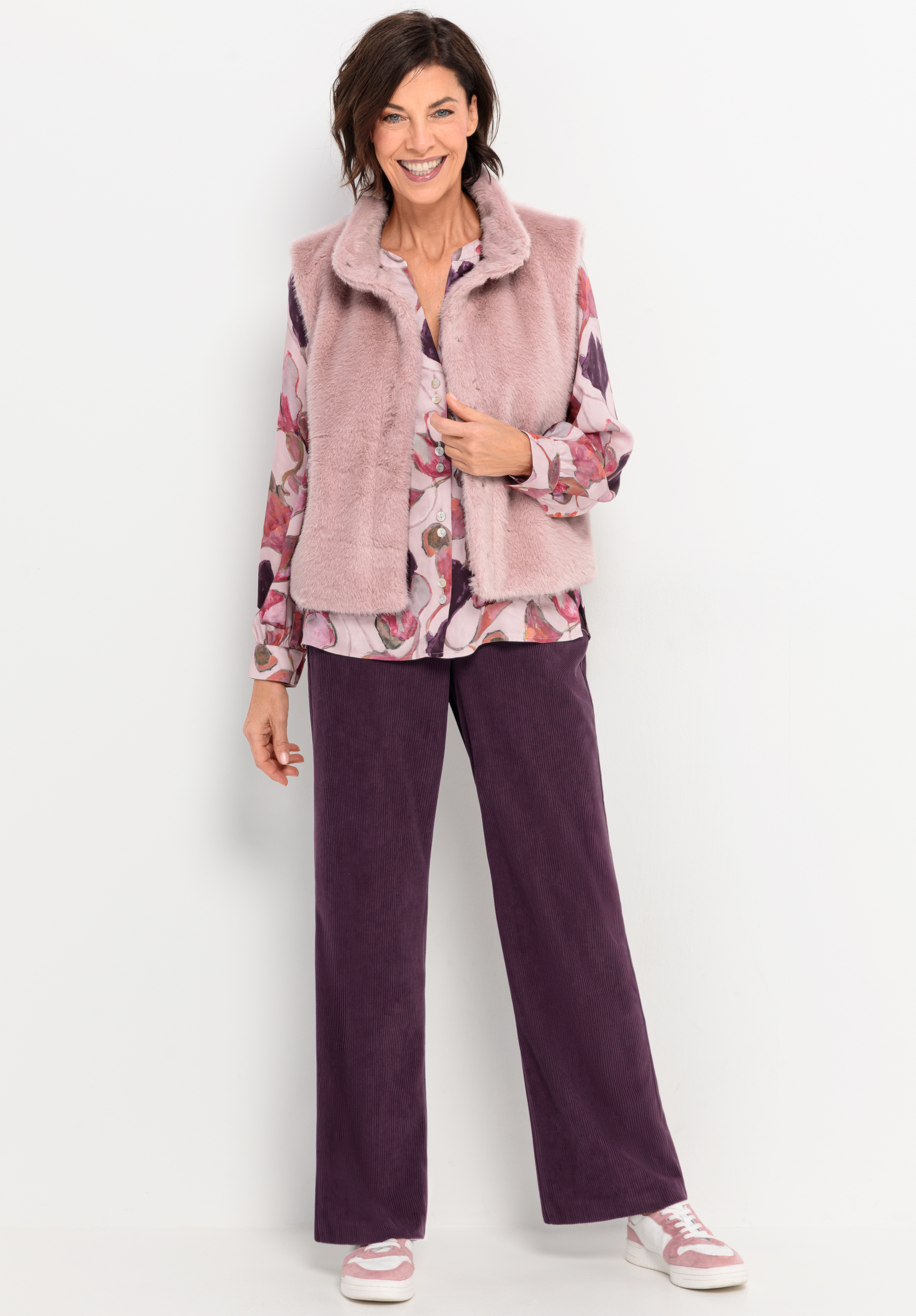 Eine Frau lächelt, während sie eine hellrosa Kunstpelzweste über einer geblümten Bluse trägt, die mit einer lilafarbenen Hose mit weitem Bein und rosa-weißen Turnschuhen kombiniert ist, während sie vor einem schlichten weißen Hintergrund steht.