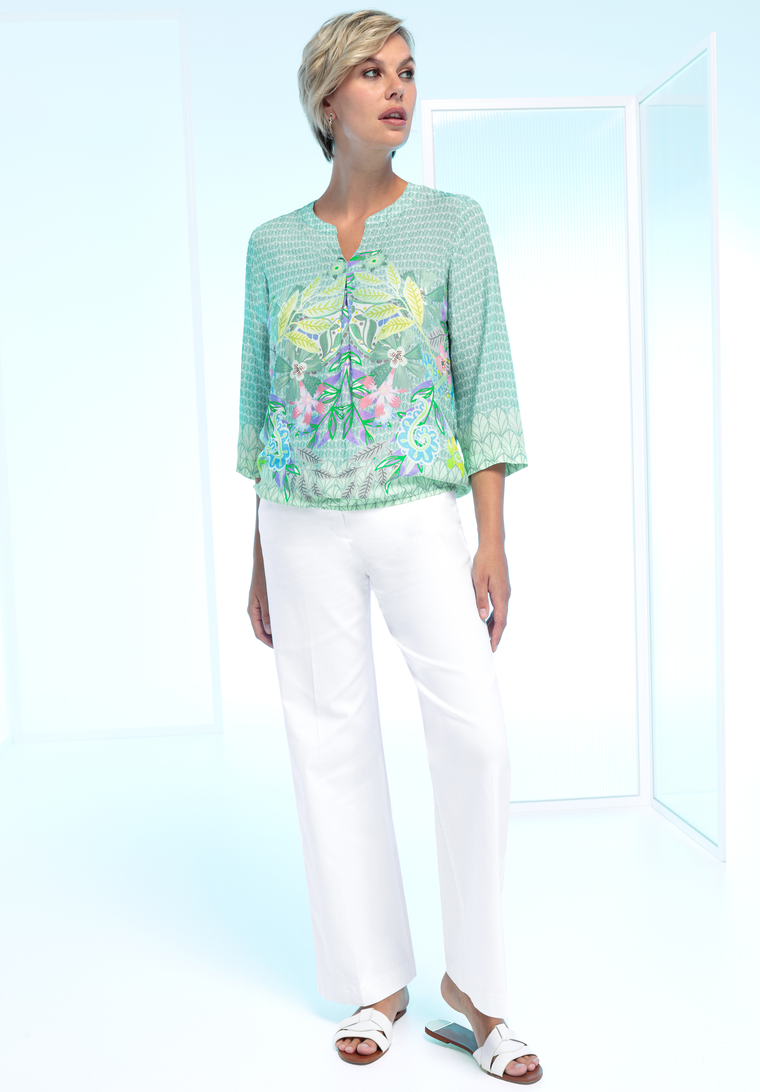 Eine Frau steht im Innenbereich und trägt eine hellgrüne Bluse mit Blumenmuster, eine weiße Hose mit geradem Bein und weiße Sandalen. Sie hat kurze blonde Haare und blickt zur Seite. Der Hintergrund ist hell und minimalistisch.