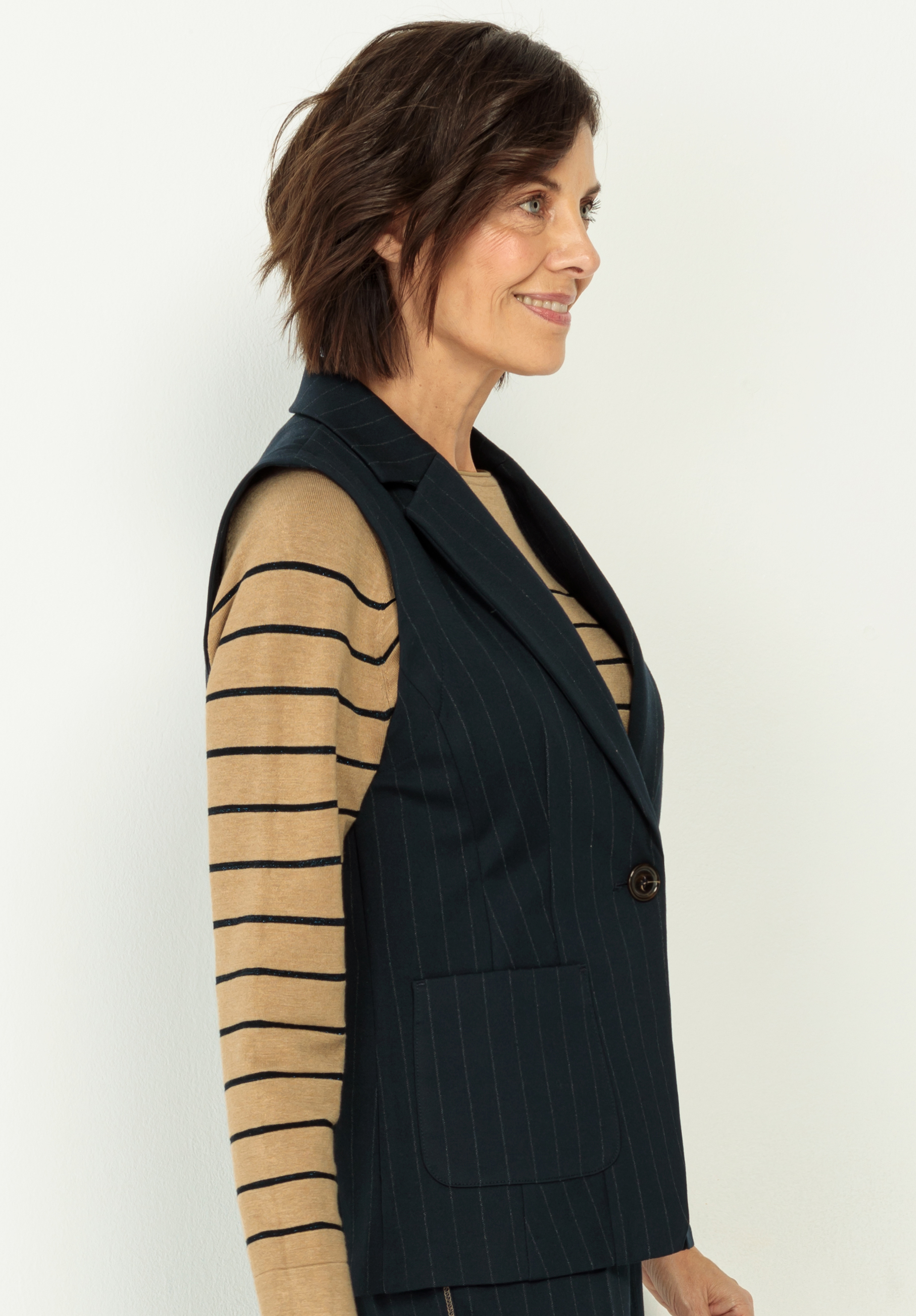 Frau mit kurzen braunen Haaren, die einen schwarzen ärmellosen Blazer mit Nadelstreifen über einem hellbraunen langärmeligen Hemd mit schwarzen Querstreifen trägt, steht im Profil vor einem schlichten hellen Hintergrund.