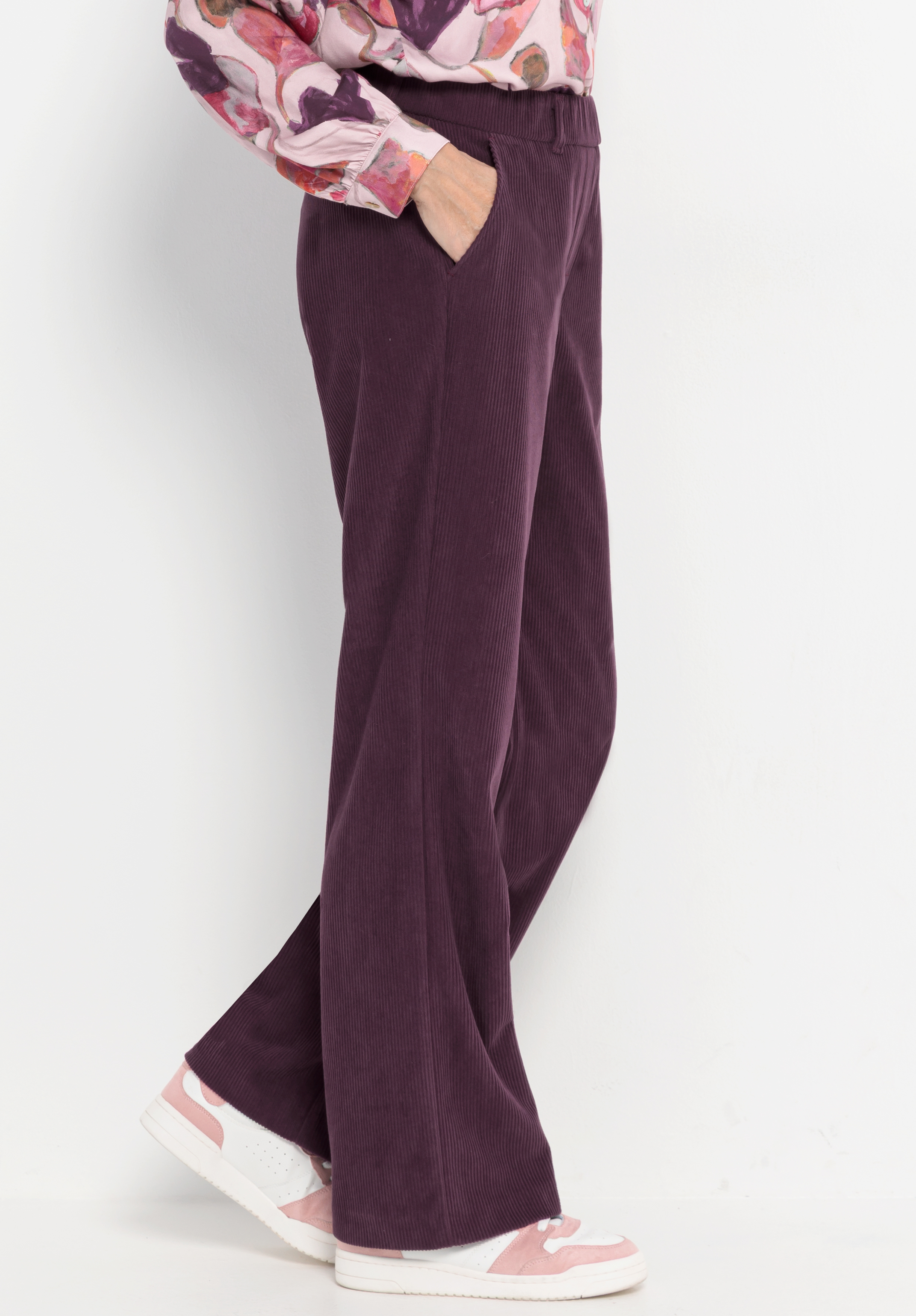Eine Person, die eine lila Cordhose mit weitem Bein und hoher Taille, eine bunt gemusterte Bluse und rosa-weiße Turnschuhe trägt, steht mit einer Hand in der Tasche vor einem weißen Hintergrund.