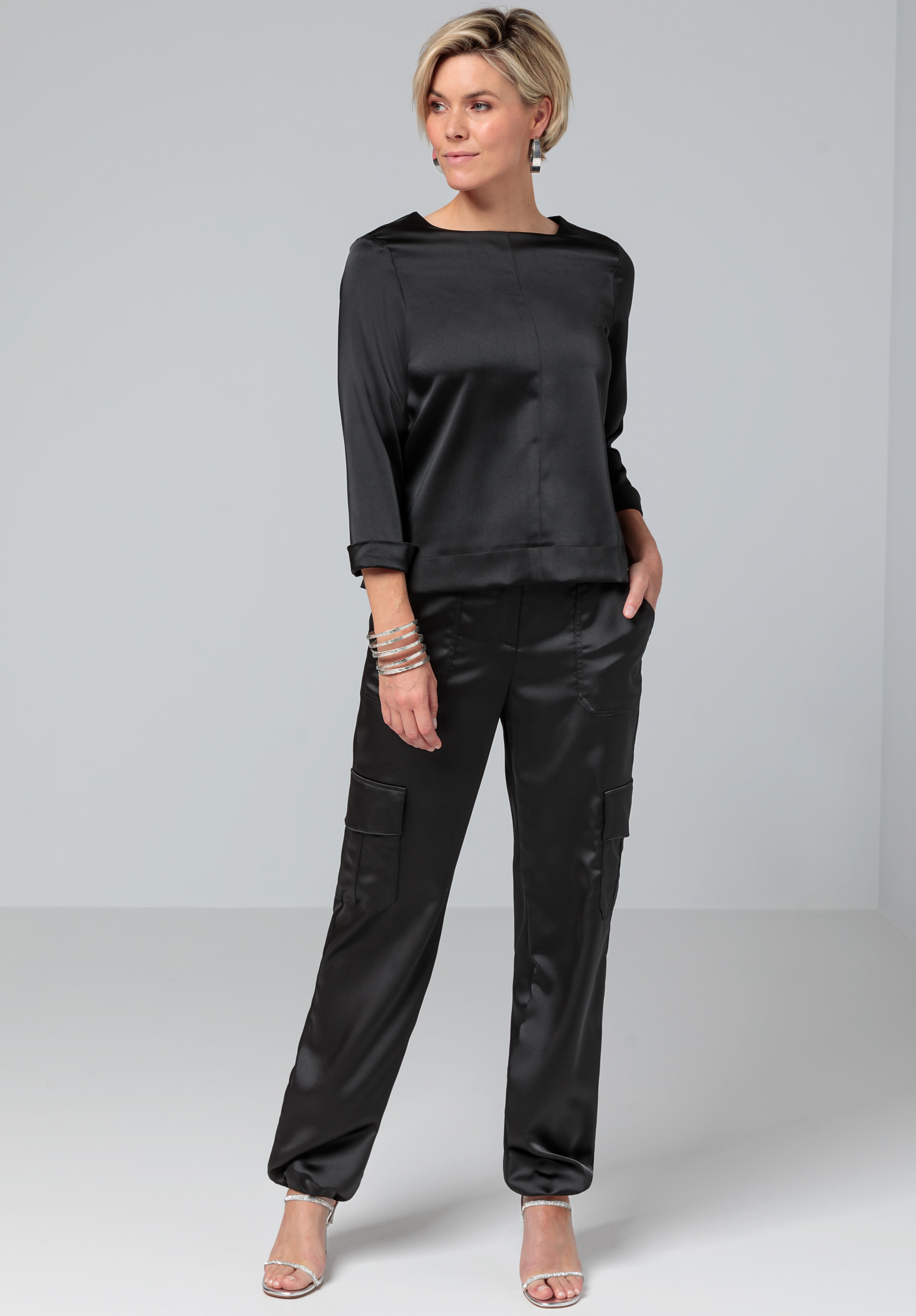 Eine Frau in einem schwarzen Satin-Langarmshirt und einer dazu passenden schwarzen Satinhose steht vor einem schlichten Hintergrund. Sie hat eine Hand in der Tasche, die andere hängt an ihrer Seite. Sie trägt silberne High Heels und Armbänder.
