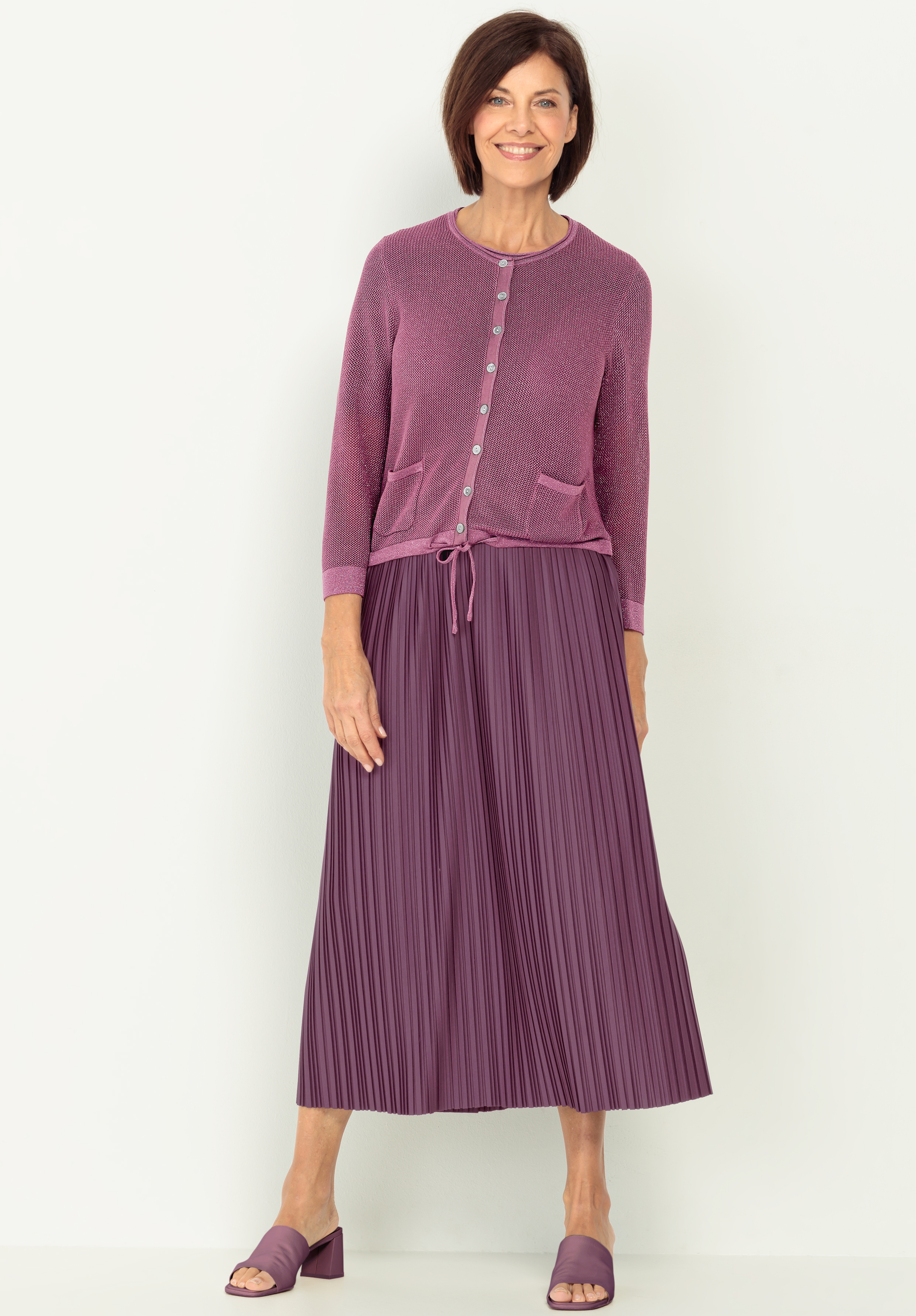 Eine Frau trägt eine hellviolette Strickjacke mit Vordertaschen und Knöpfen, gepaart mit einem passenden plissierten Midirock und offenen Schuhen mit Blockabsatz. Sie hat kurzes braunes Haar.