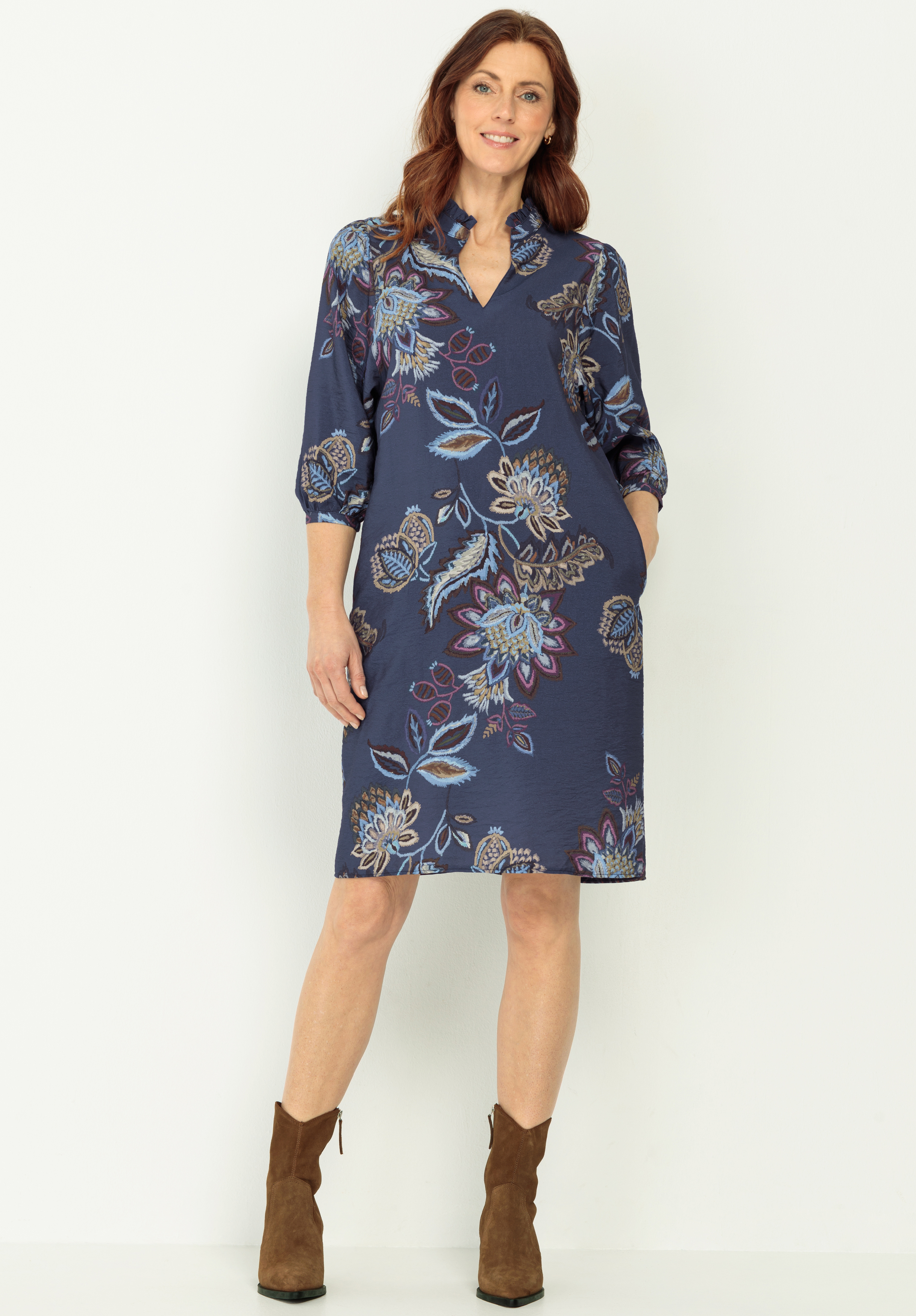 Eine Frau steht vor einem hellen Hintergrund und trägt ein knielanges, langärmeliges, marineblaues Kleid mit buntem Blumenmuster und braune Stiefeletten. Sie lächelt und hat die Hände in den Taschen.