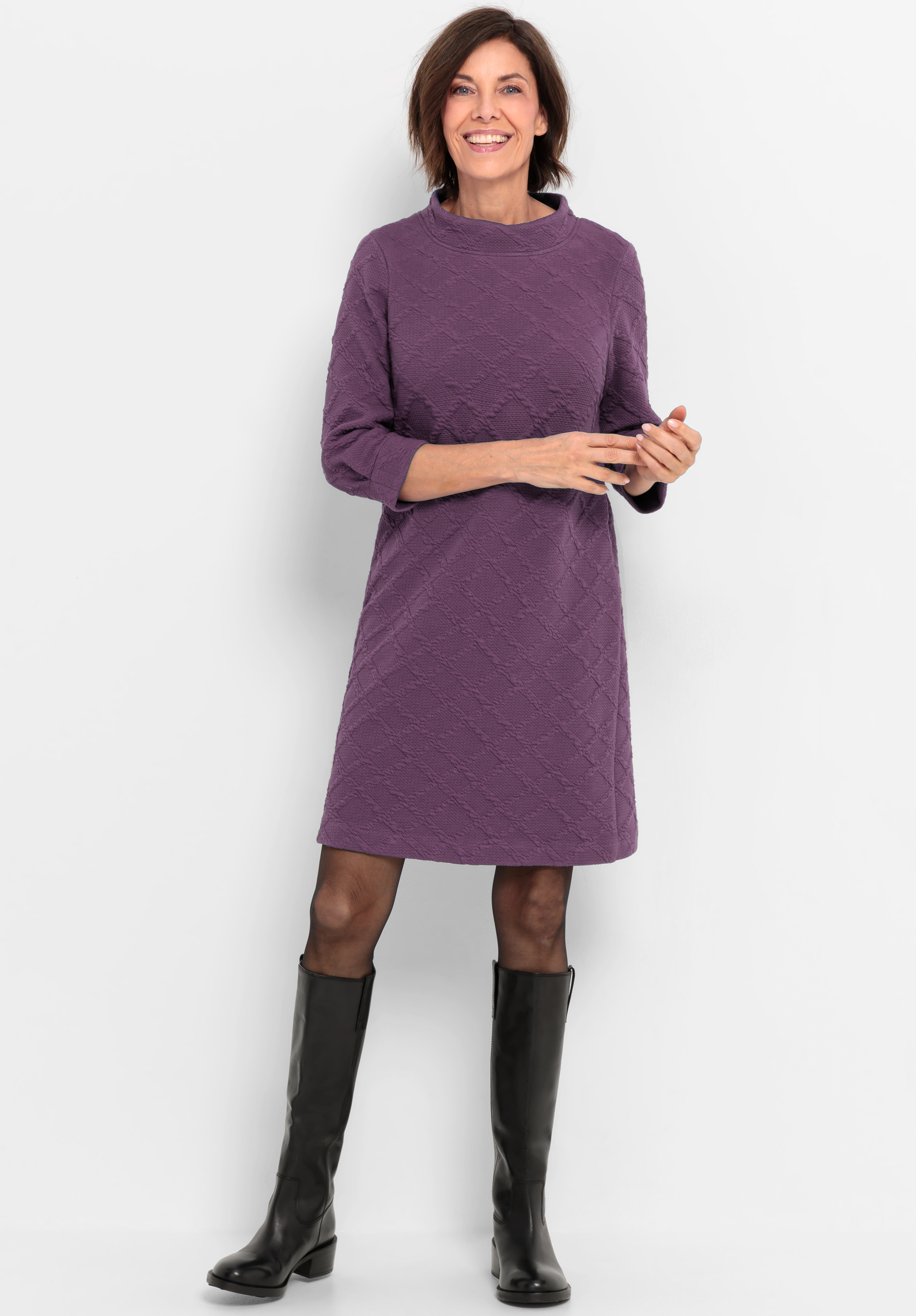 Eine Frau steht lächelnd vor einem weißen Hintergrund und trägt ein knielanges, langärmeliges lilafarbenes Kleid mit einem strukturierten Muster, eine durchsichtige schwarze Strumpfhose und schwarze kniehohe Stiefel.