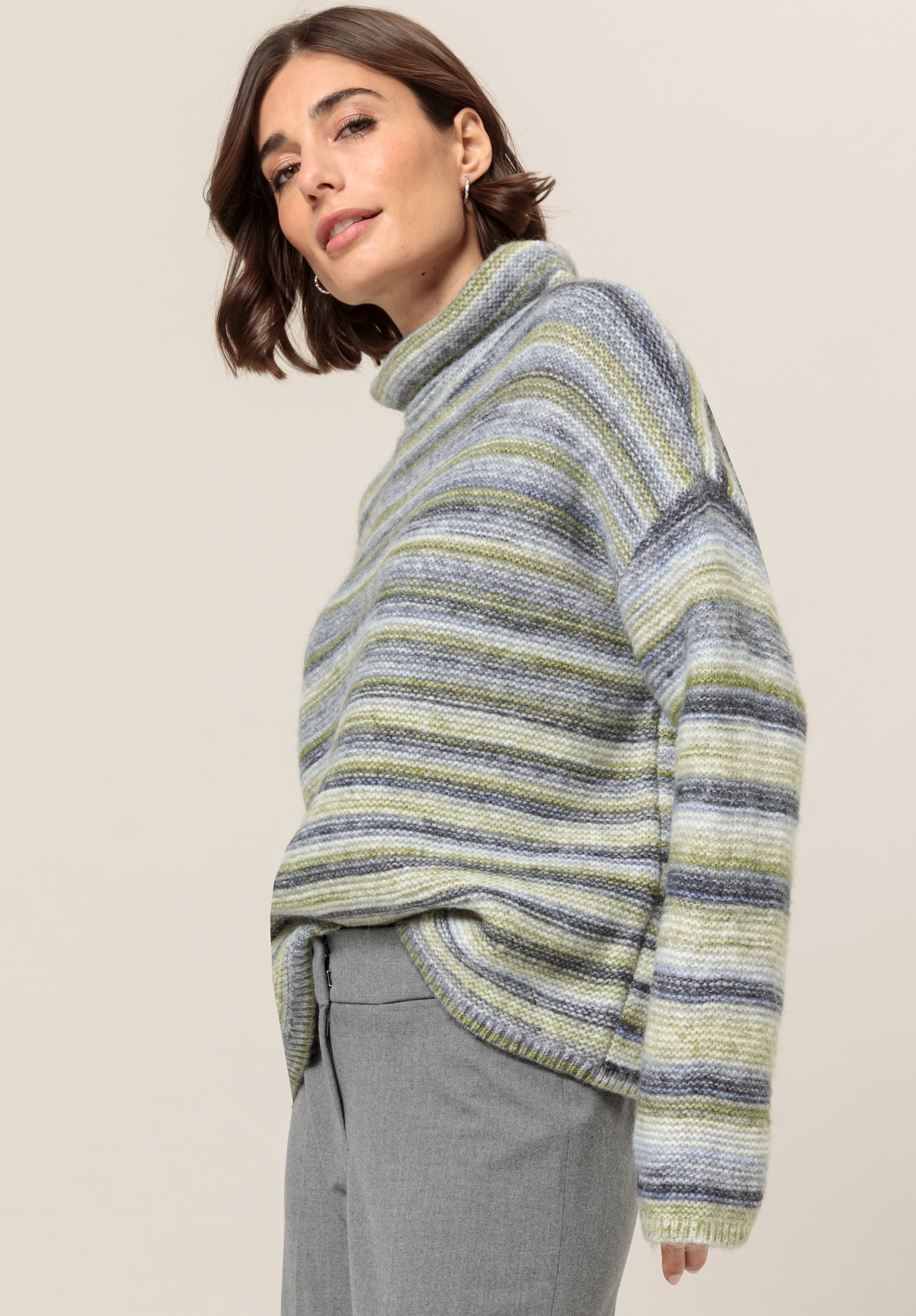 Eine Frau mit kurzen braunen Haaren trägt einen lockeren, gestreiften Rollkragenpullover in pastellfarbenen Grün-, Grau- und Blautönen, kombiniert mit hellgrauen Hosen, und steht vor einem schlichten beigen Hintergrund.