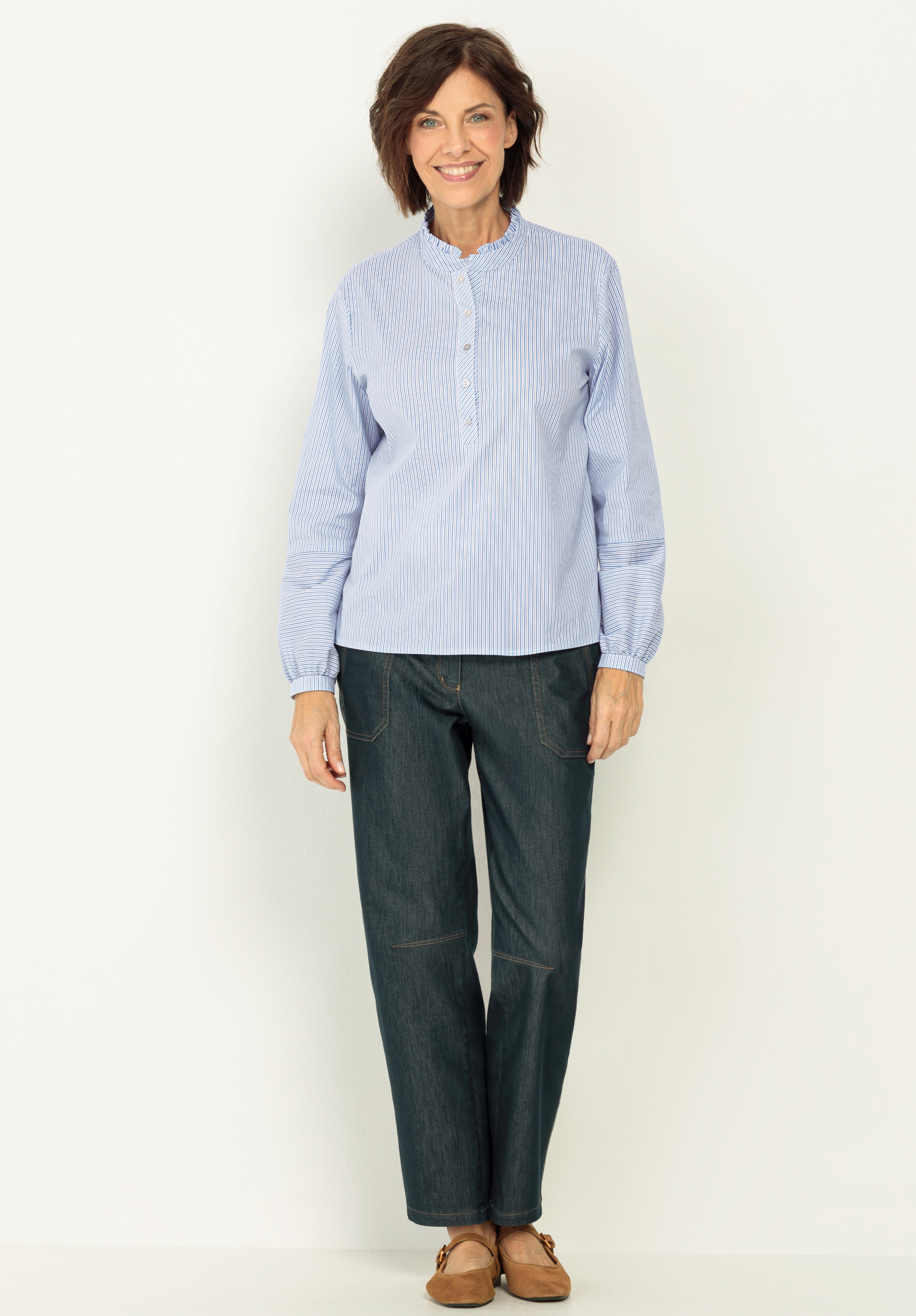 Eine Frau mit kurzen braunen Haaren steht lächelnd vor einem weißen Hintergrund. Sie trägt ein hellblau gestreiftes Button-up-Hemd, eine weite dunkle Hose und braune Schuhe.