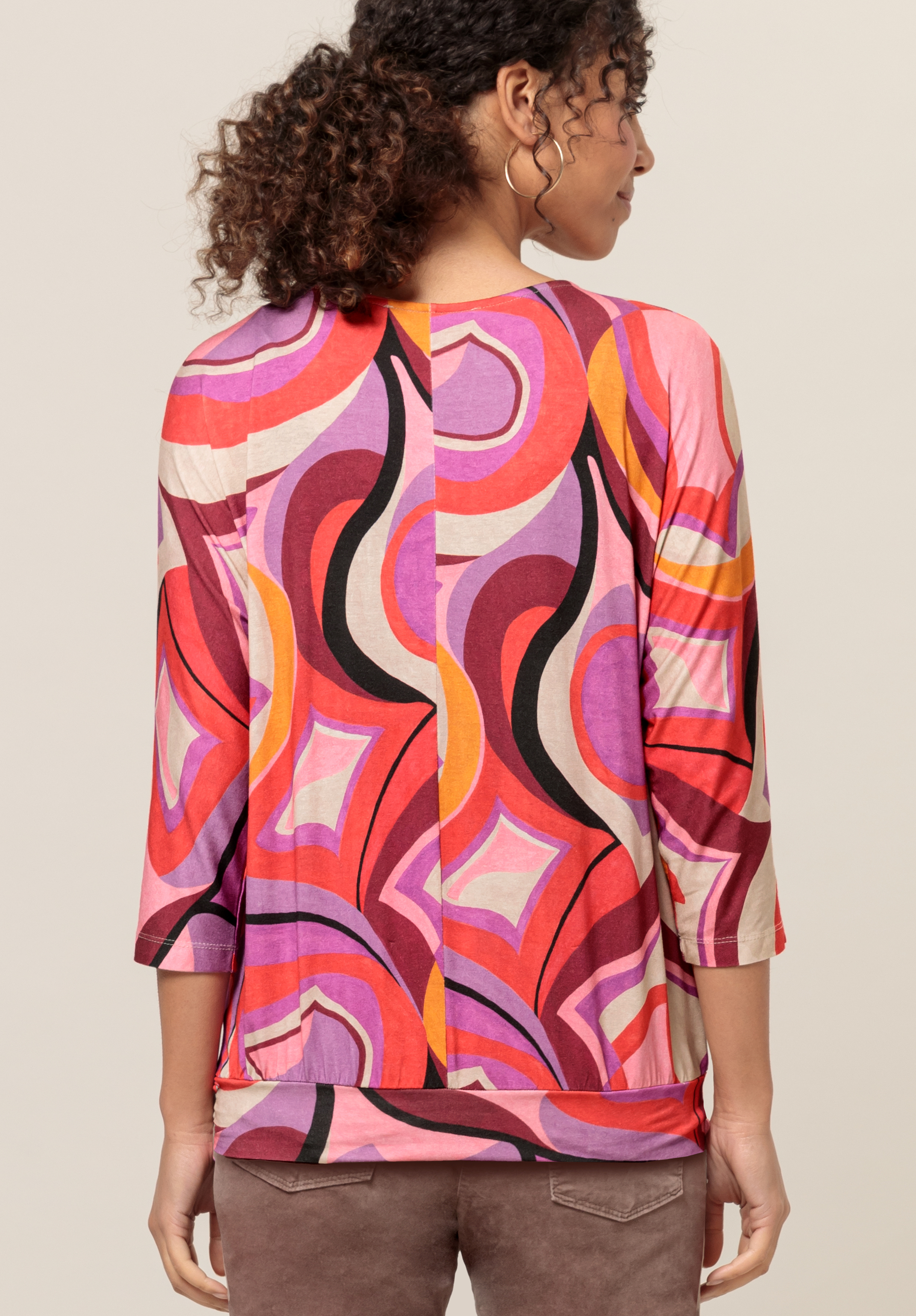 Eine Frau mit lockigem Haar, von hinten gesehen, trägt ein farbenfrohes Langarmshirt mit einem auffälligen, abstrakten Muster in Rosa-, Rot-, Lila- und Orangetönen. Sie steht vor einem schlichten, hellen Hintergrund.