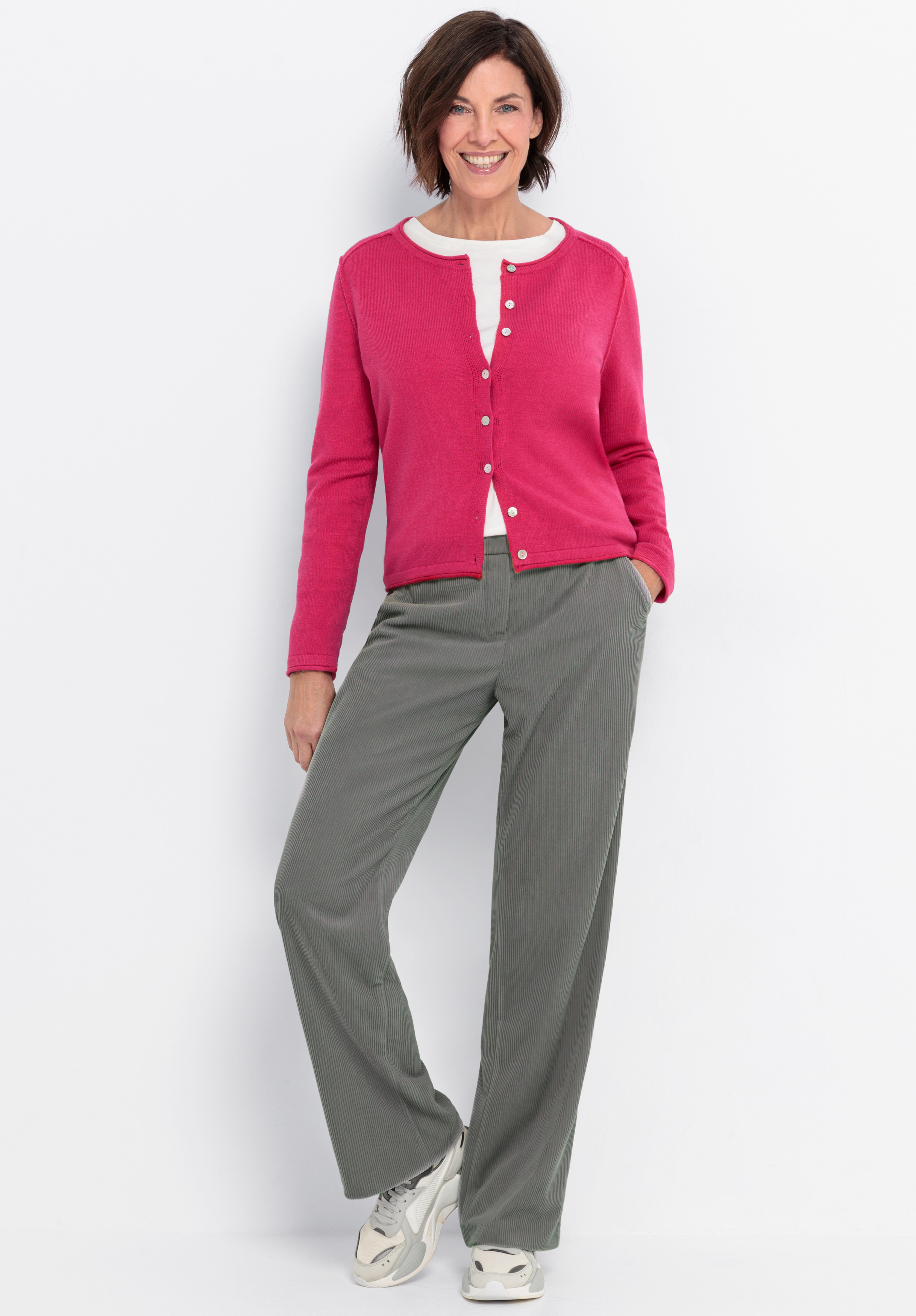 Eine Frau mit kurzen braunen Haaren lächelt, während sie eine hellrosa Strickjacke, ein weißes Oberteil, eine graue Hose mit weitem Bein und weiße Turnschuhe trägt. Sie steht vor einem schlichten weißen Hintergrund und hat eine Hand in der Tasche.