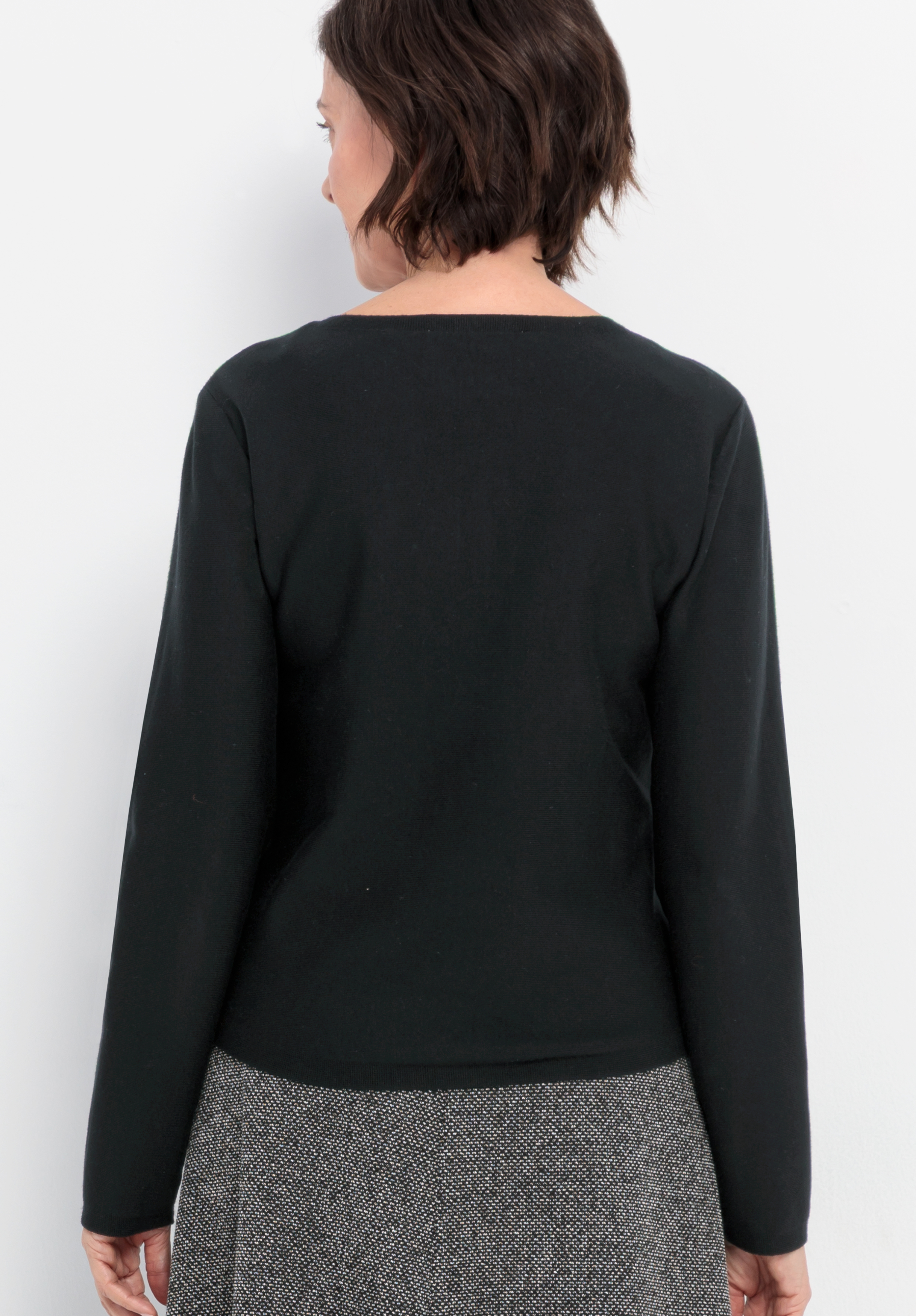 Eine Frau mit kurzen braunen Haaren ist von hinten zu sehen; sie trägt ein schwarzes Langarm-Oberteil und einen grauen Rock mit strukturiertem Muster und steht vor einem schlichten weißen Hintergrund.