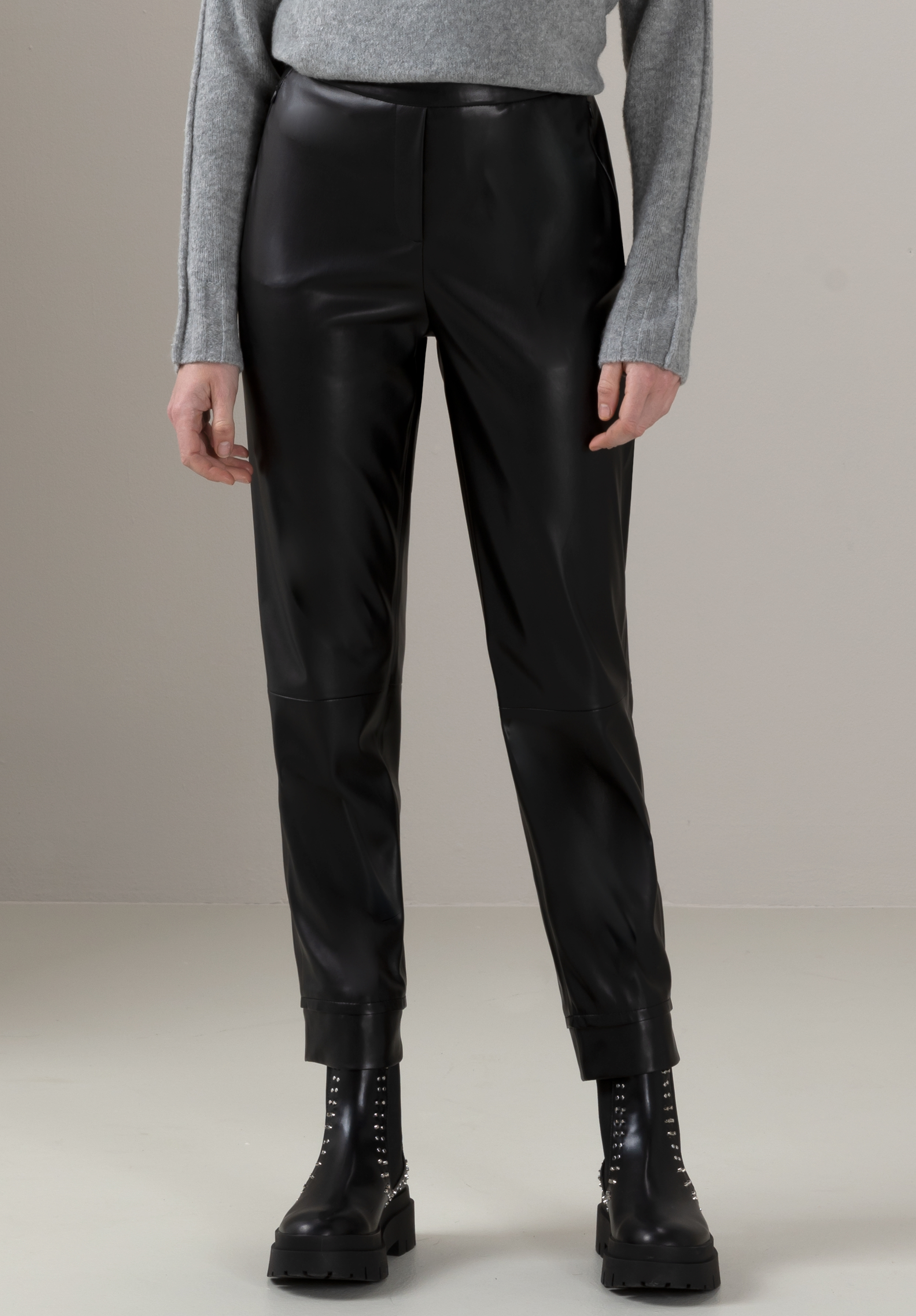Eine Person, die eine schwarze Kunstlederhose, einen hellgrauen Langarmpullover und schwarze Stiefeletten mit Nieten trägt, steht vor einem schlichten hellen Hintergrund.
