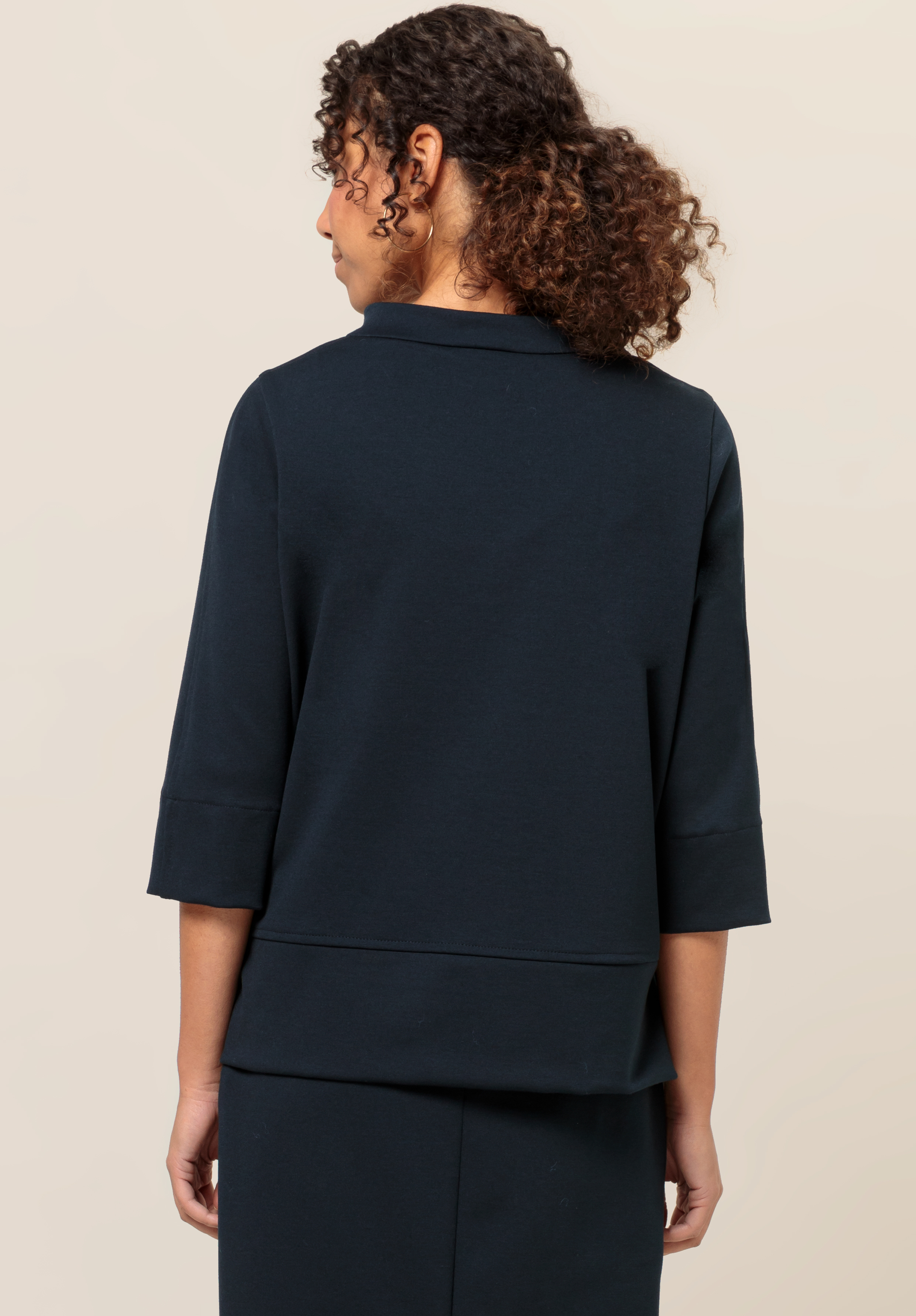 Eine Frau mit lockigem Haar blickt abgewandt. Sie trägt ein dunkles Oberteil mit Dreiviertelärmeln und einen dazu passenden Rock. Der Hintergrund ist schlicht und hell.