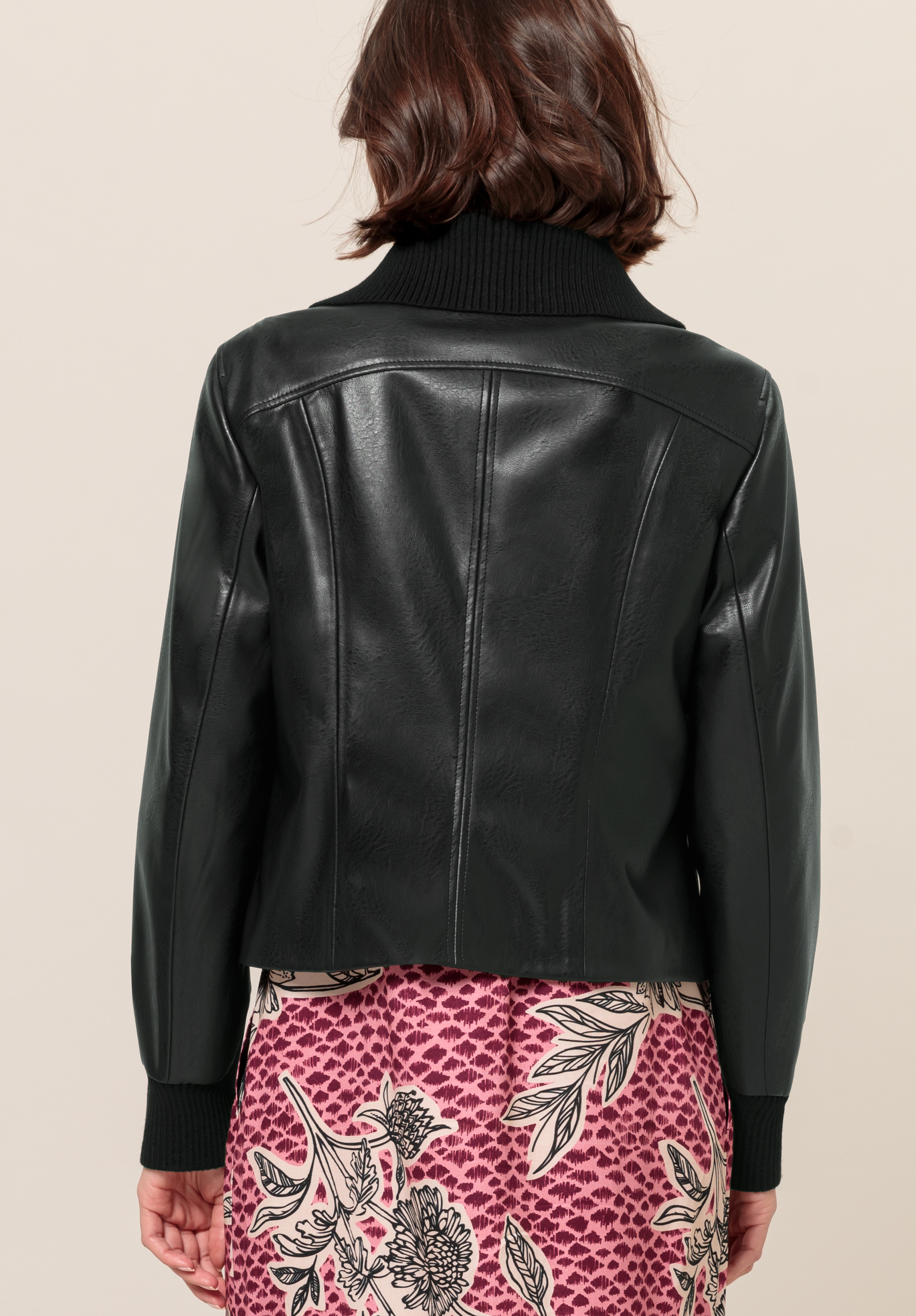 Eine Frau mit kurzen braunen Haaren, von hinten gesehen, trägt eine schwarze Lederjacke mit geripptem Strickkragen und Manschetten über einem rosa gemusterten Kleid mit Blumenmuster.