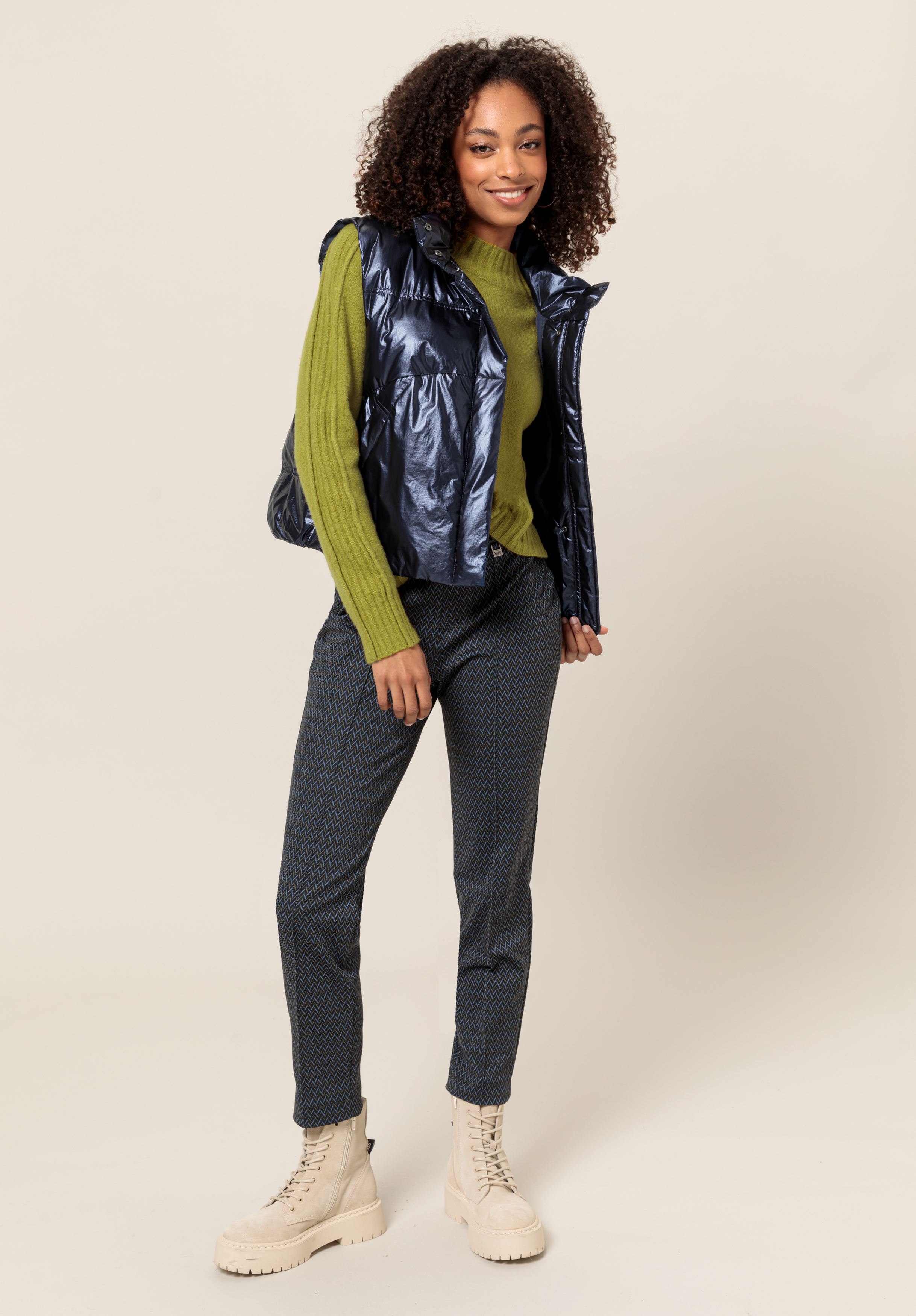 Eine Frau mit lockigem Haar lächelt, während sie eine glänzende marineblaue Weste über einem grünen Pullover, gemusterte dunkle Hosen und cremefarbene Schnürstiefel trägt und vor einem schlichten hellen Hintergrund steht.