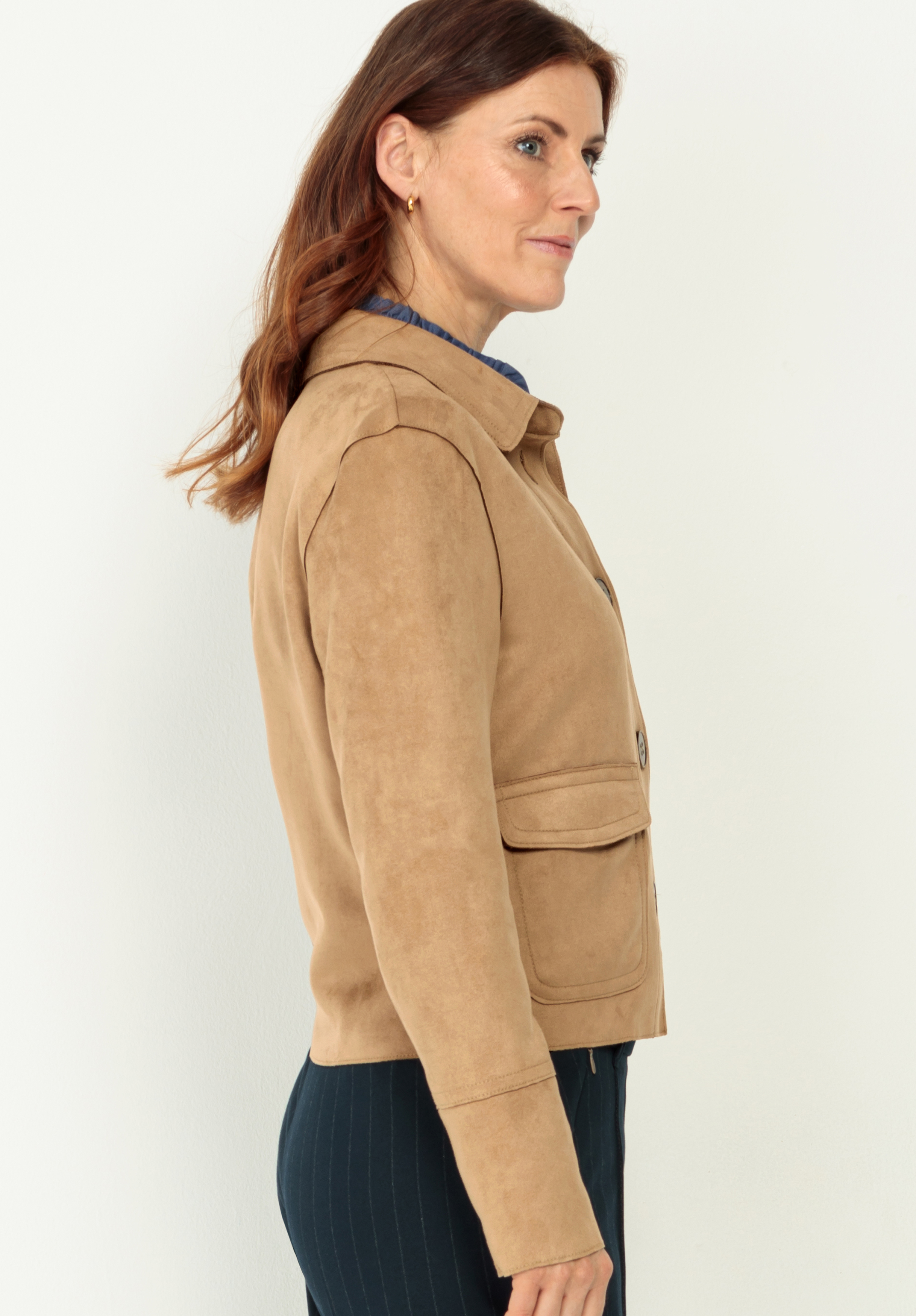 Eine Frau mit langem kastanienbraunem Haar, die eine hellbraune Wildlederjacke und eine dunkle Hose trägt, steht seitlich mit Blick nach links vor einem schlichten hellen Hintergrund.