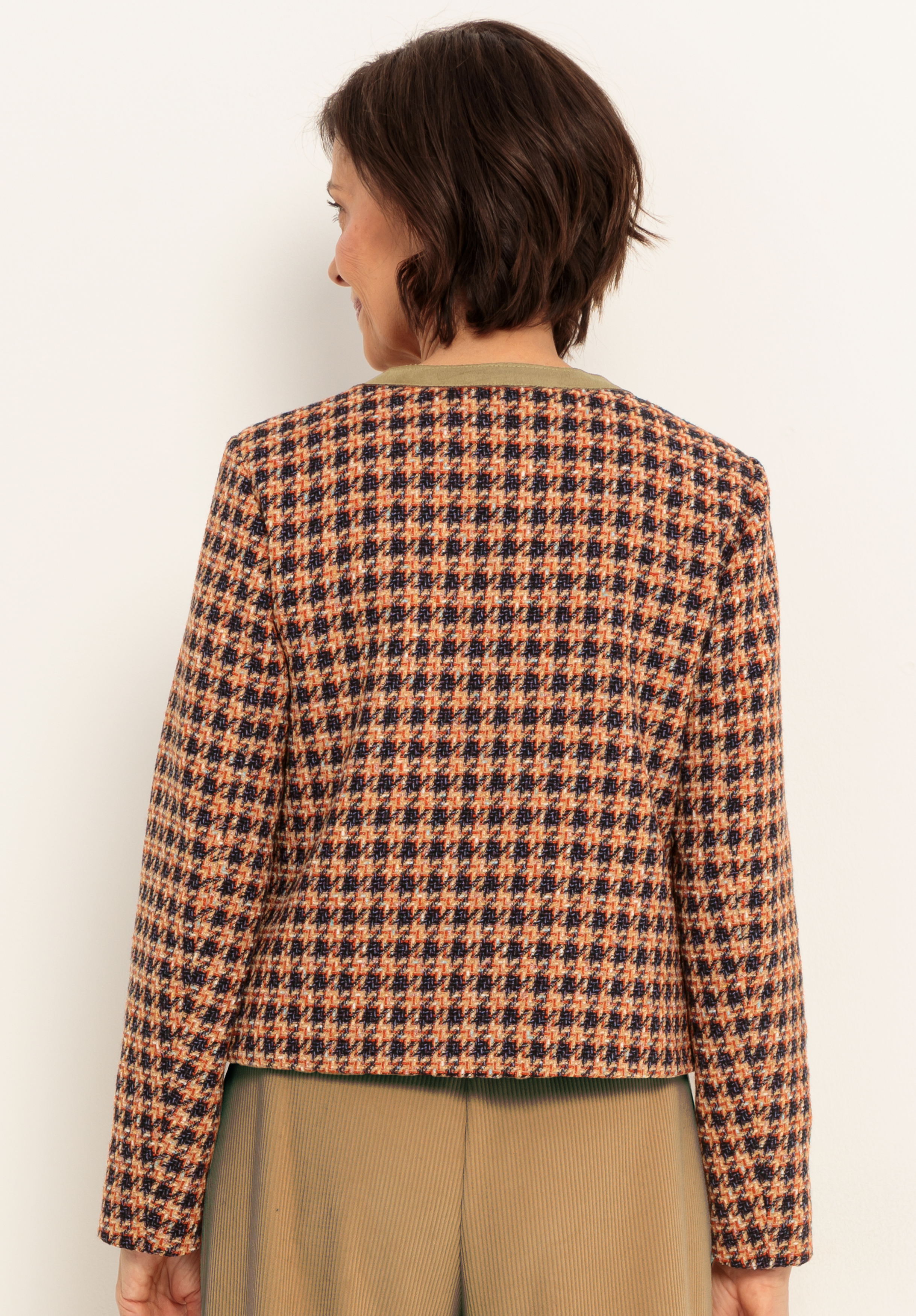 Eine Frau steht mit dem Gesicht zur Kamera und trägt eine langärmelige, mehrfarbige Jacke mit Hahnentrittmuster und eine helle Bundfaltenhose. Ihr kurzes, dunkles Haar ist zu lockeren Wellen gestylt.