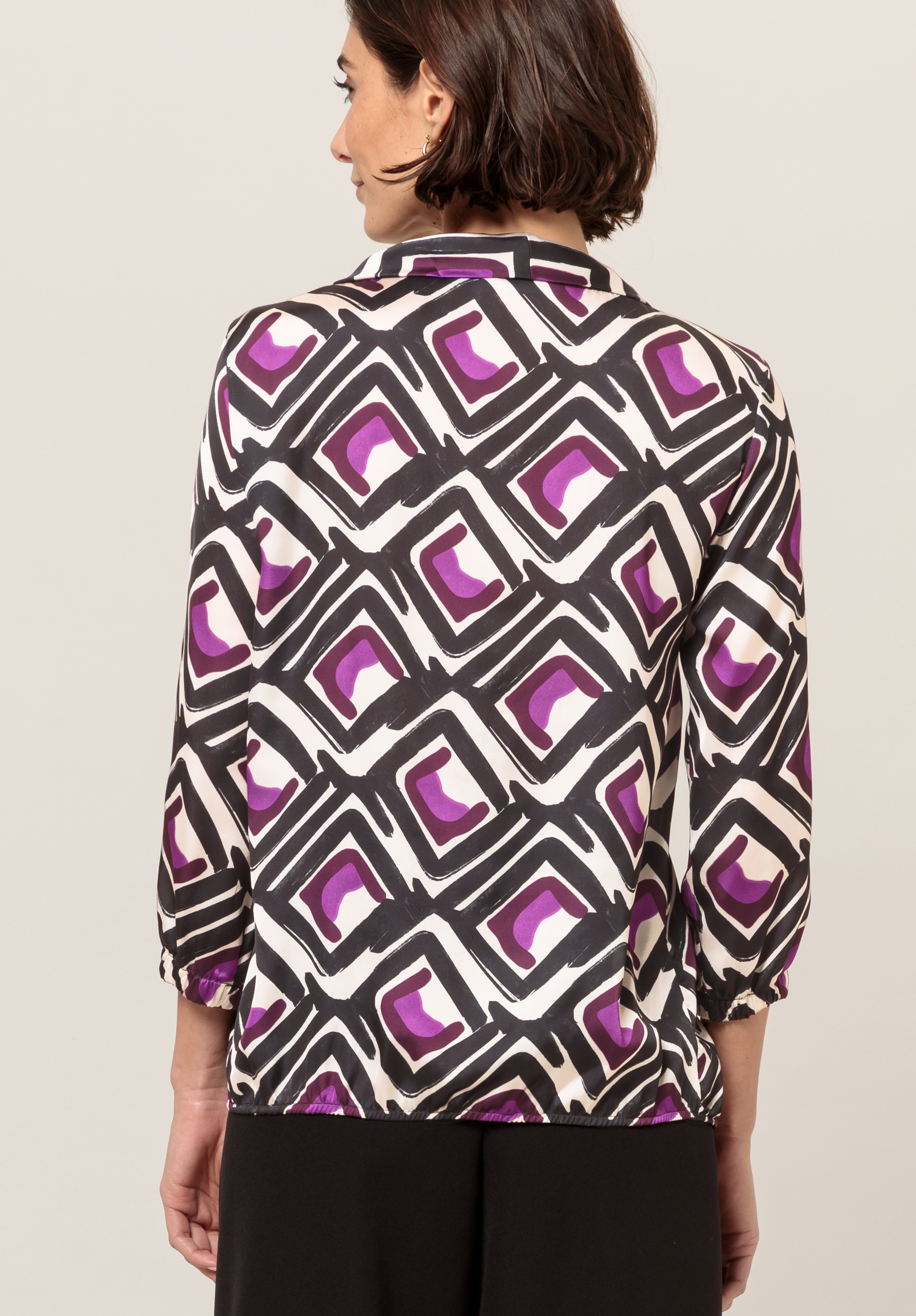 Von hinten ist eine Frau mit kurzen braunen Haaren zu sehen. Sie trägt eine gemusterte Bluse mit geometrischen Mustern in Schwarz, Weiß und Lila und Dreiviertelärmeln. Außerdem trägt sie eine schwarze Hose.