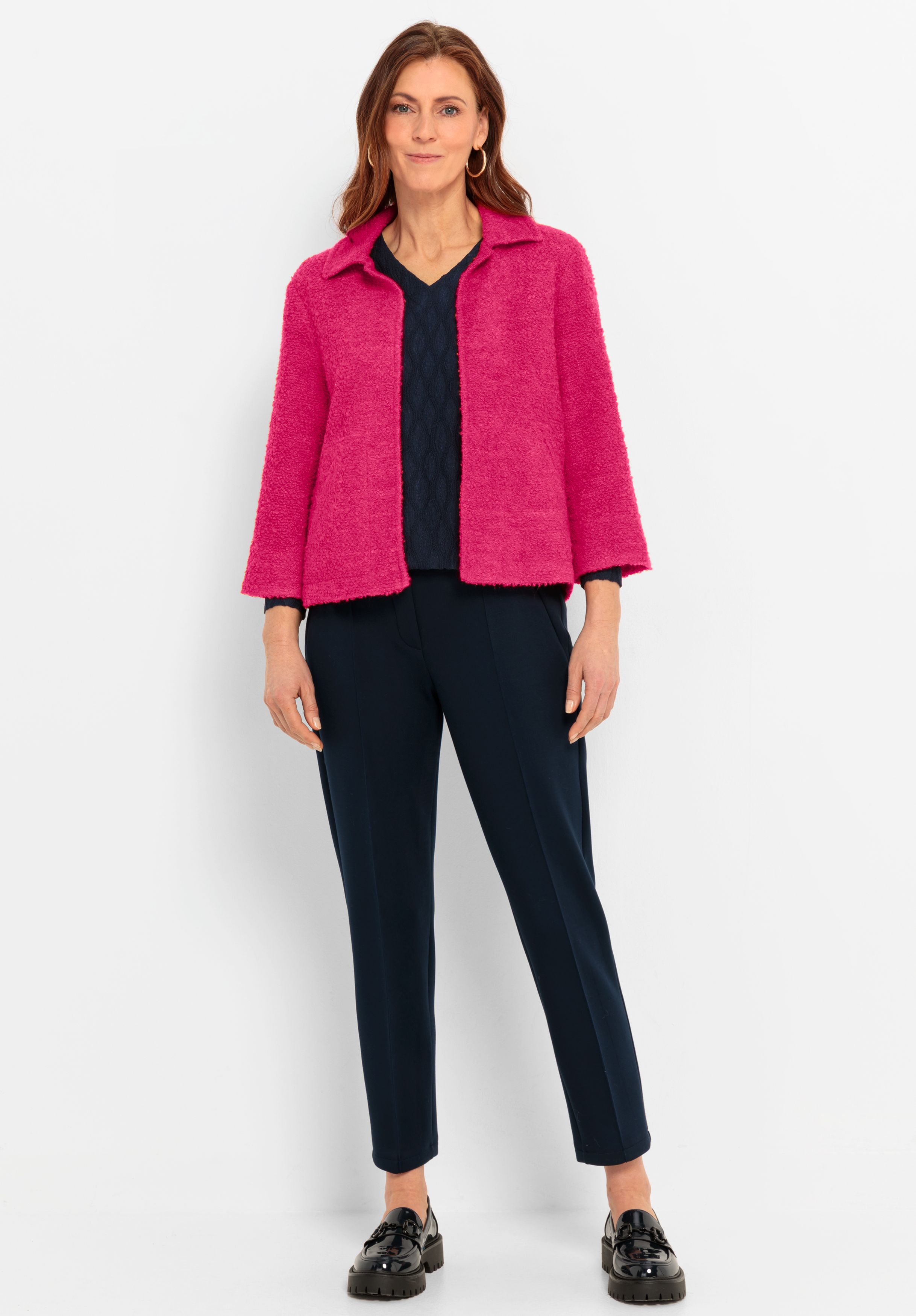 Eine Frau steht vor einem weißen Hintergrund und trägt eine hellrosa Jacke, ein marineblaues strukturiertes Oberteil, eine marineblaue Slim-Fit-Hose und schwarze Slipper. Sie hat schulterlanges braunes Haar und lächelt leicht.