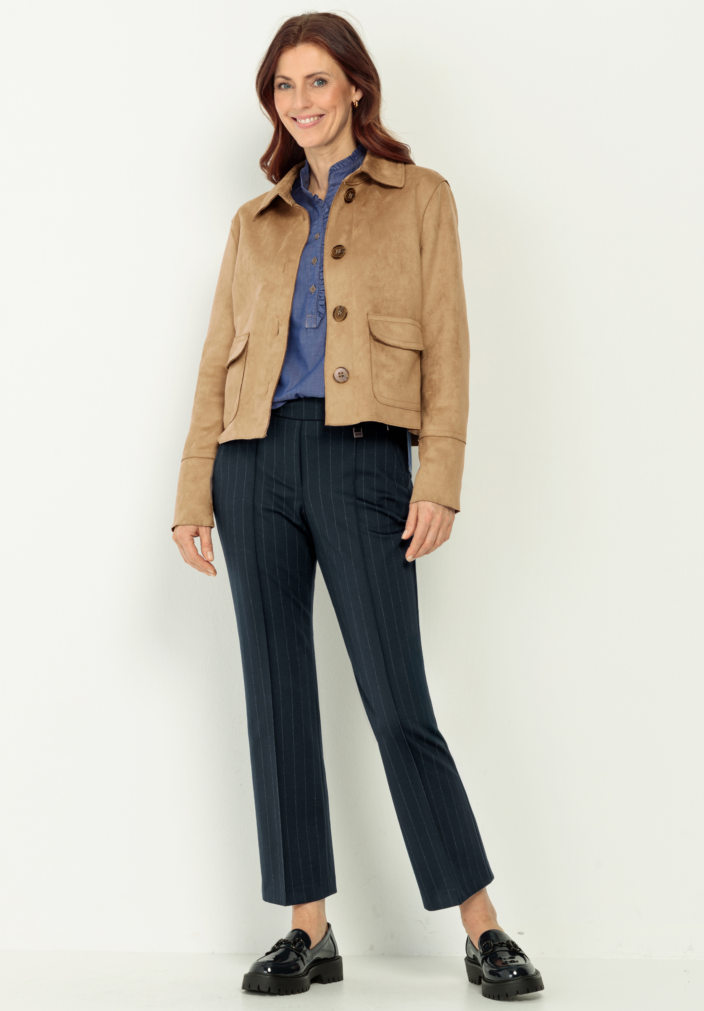 Eine Frau steht lächelnd vor einem hellen Hintergrund. Sie trägt eine hellbraune Jacke über einem blauen Button-up-Hemd, eine dunkle Nadelstreifenhose und schwarz glänzende Slipper.