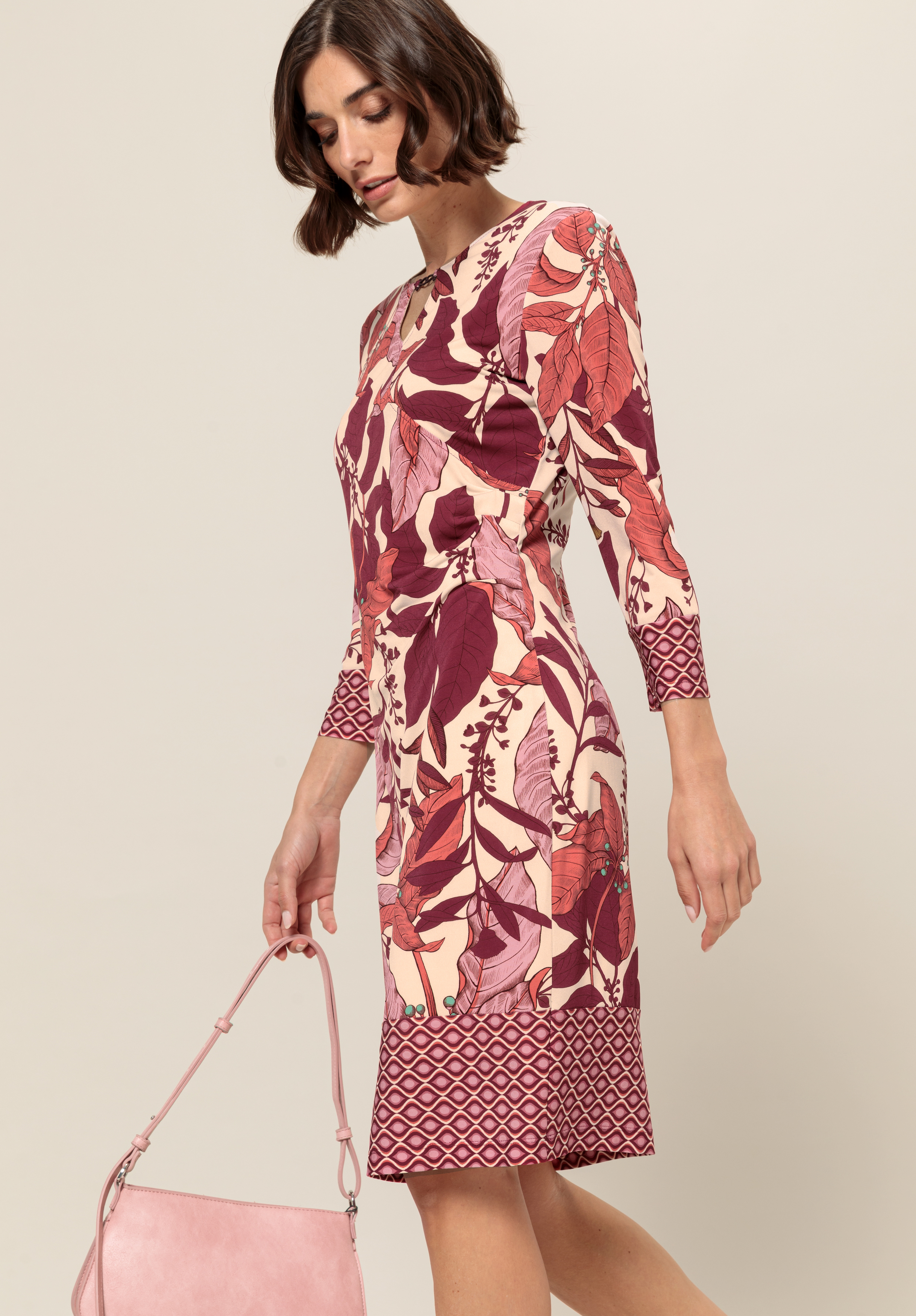 Eine Frau in einem figurbetonten, knielangen Kleid mit kastanienbraunen, rosa und beigen Blatt- und geometrischen Mustern hält eine blassrosa Handtasche. Sie hat kurzes braunes Haar und blickt nach unten. Sie steht vor einem schlichten hellen Hintergrund.