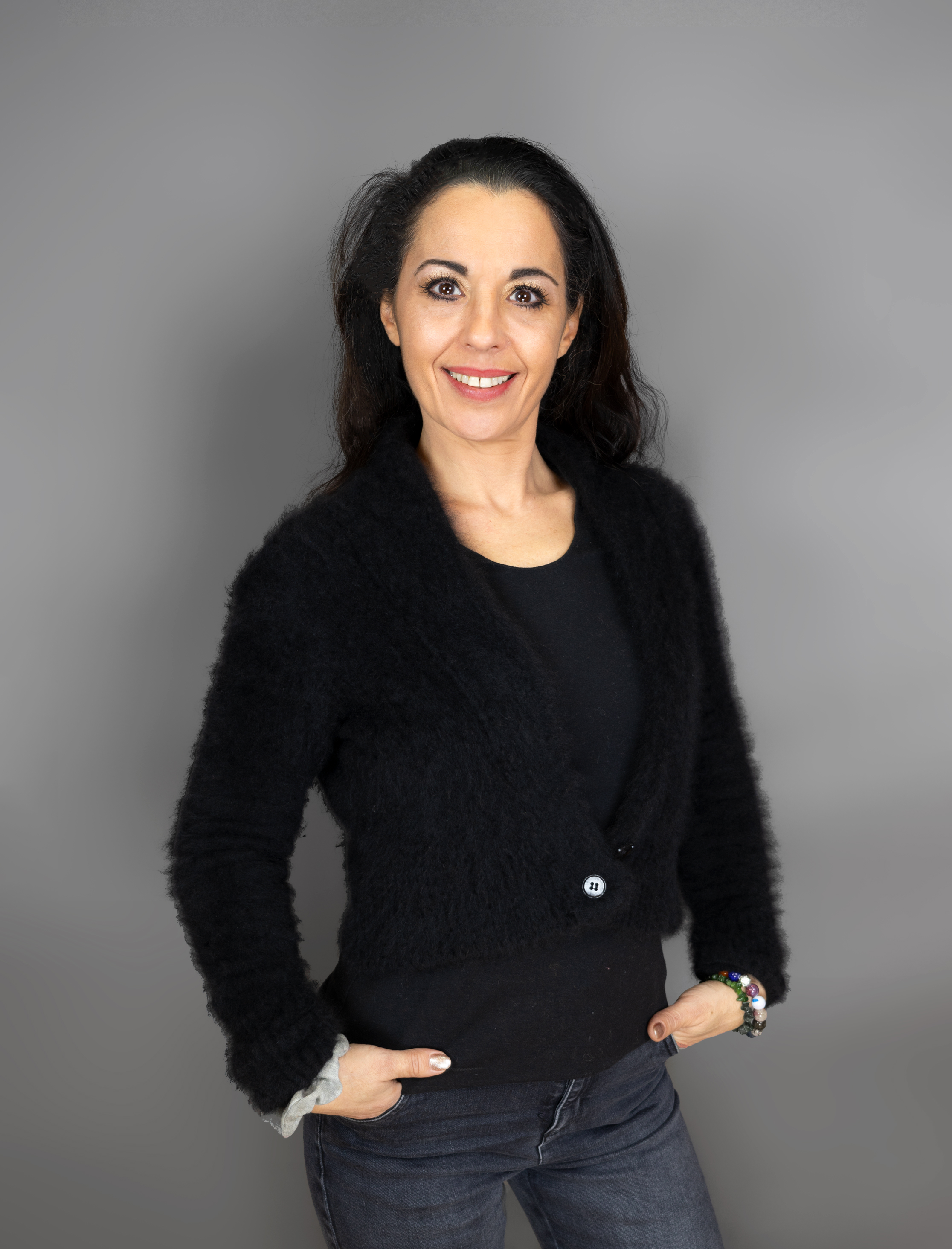 Eine Frau mit langen dunklen Haaren posiert vor einem grauen Hintergrund. Sie trägt eine schwarze, flauschige Strickjacke über einem schwarzen Top und dunkle Jeans. Sie lächelt selbstbewusst, die linke Hand ruht auf der Hüfte, die rechte in der Tasche.