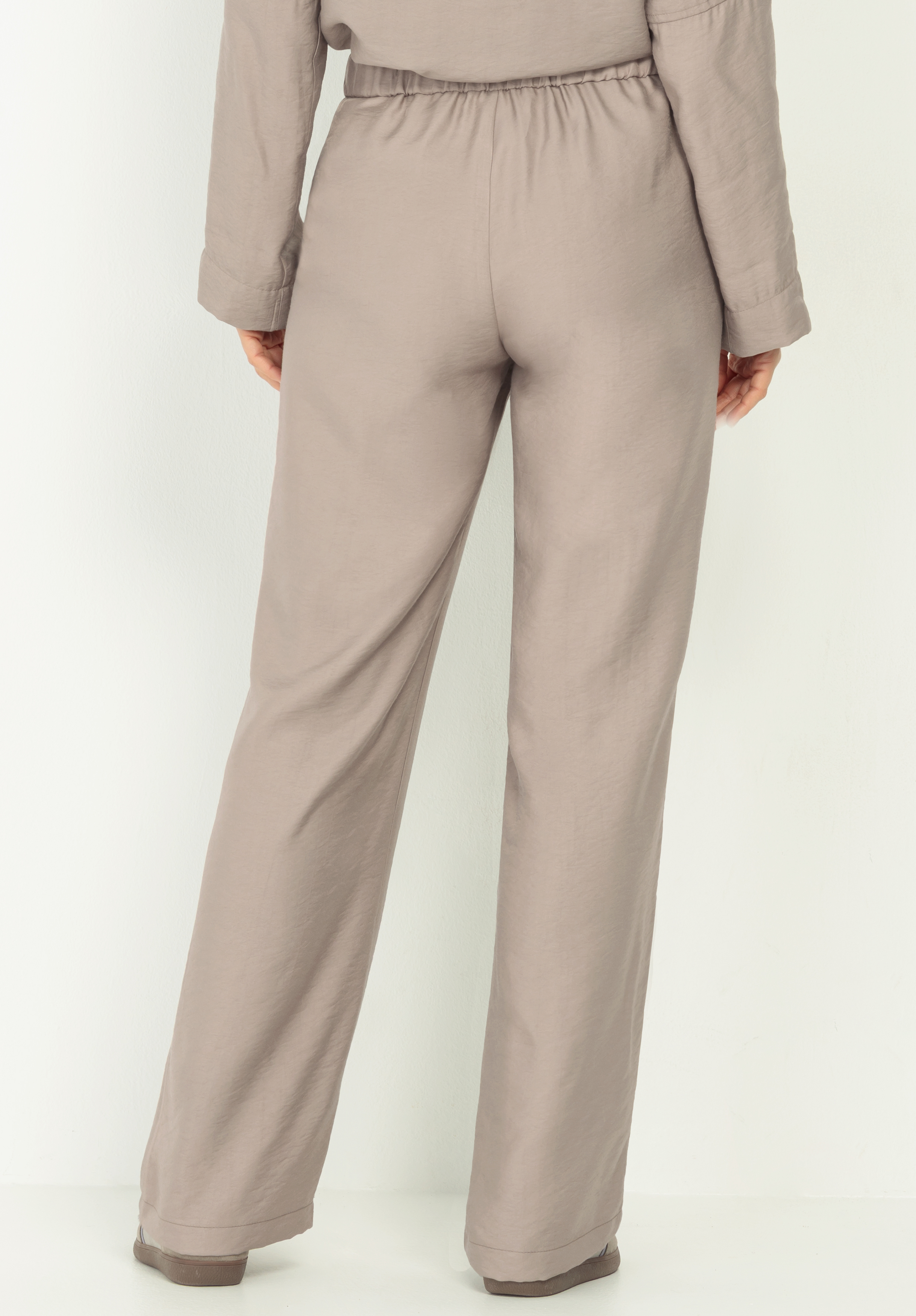 Eine Person, die eine locker sitzende, helltaupefarbene Hose und ein dazu passendes langärmeliges Oberteil trägt, steht mit dem Gesicht zur Kamera vor einem schlichten weißen Hintergrund.