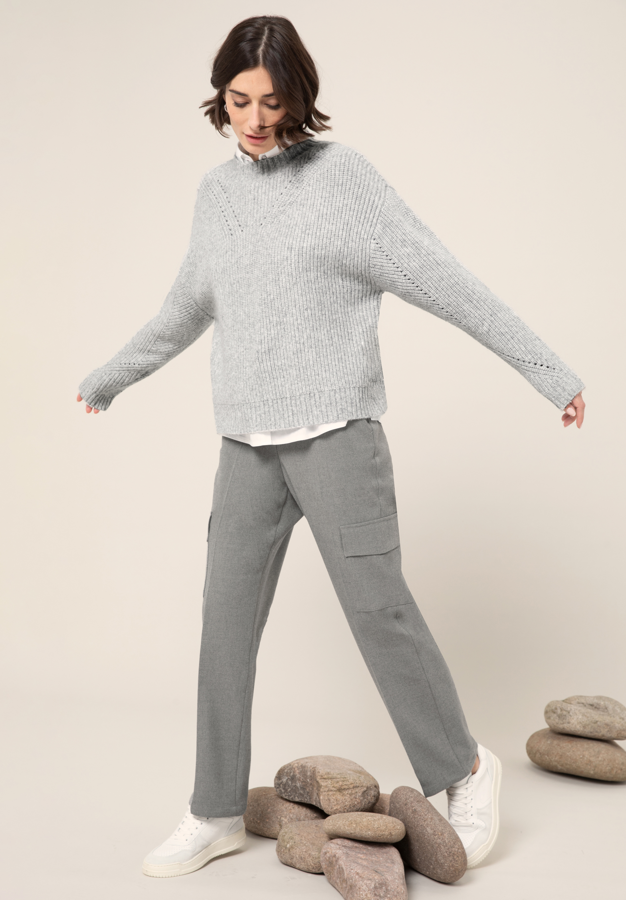Eine Frau in einem grauen Strickpullover, einem weißen Hemd, einer grauen Hose und weißen Turnschuhen steht mit ausgestreckten Armen da und balanciert auf gestapelten glatten Steinen vor einem neutralen Hintergrund.