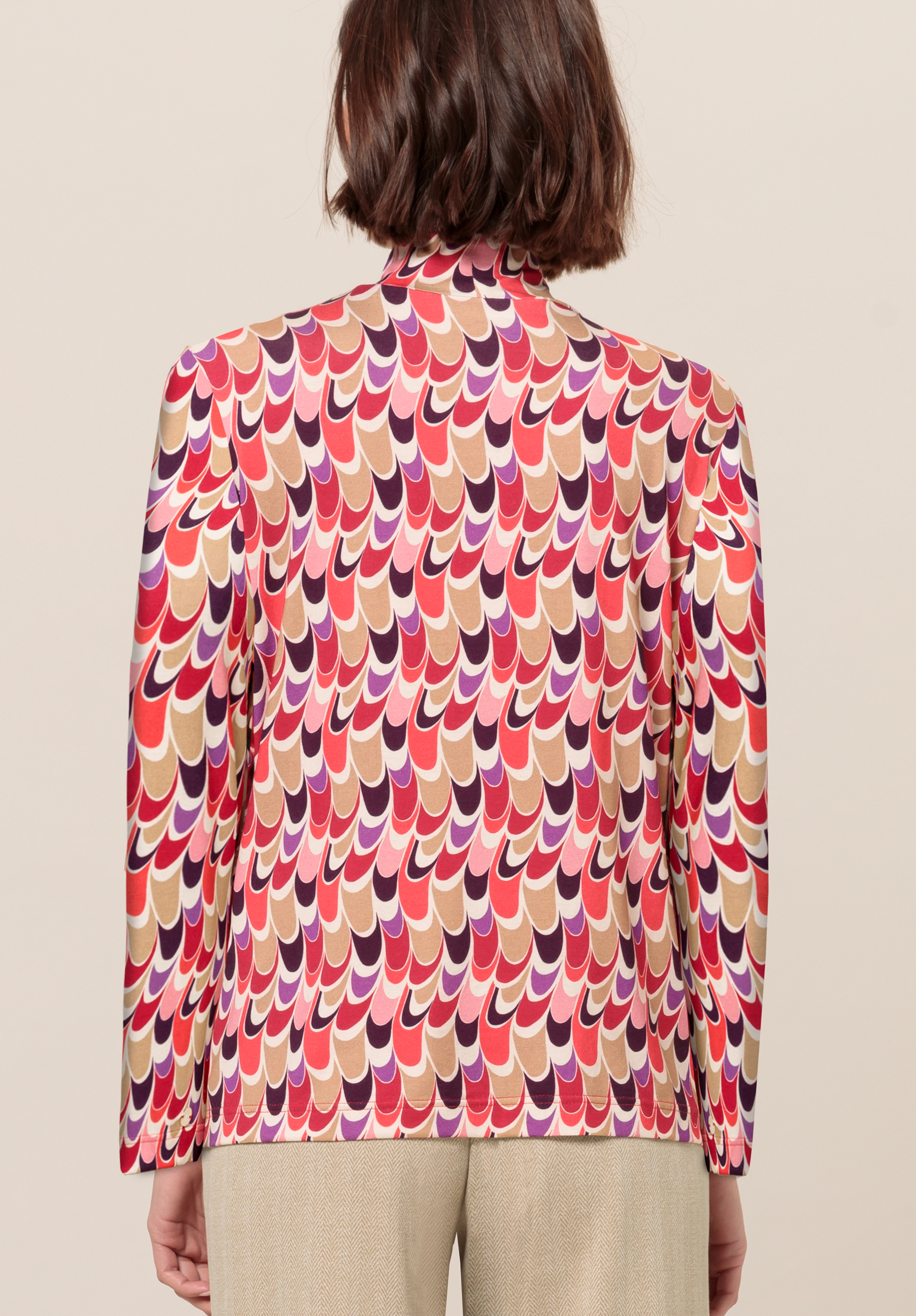 Eine Person mit schulterlangem braunem Haar ist von hinten zu sehen. Sie trägt einen Blazer mit einem kräftigen, mehrfarbigen, gewellten geometrischen Muster in Rot-, Rosa-, Lila- und Beigetönen. Dazu trägt sie eine hellbeige Hose.