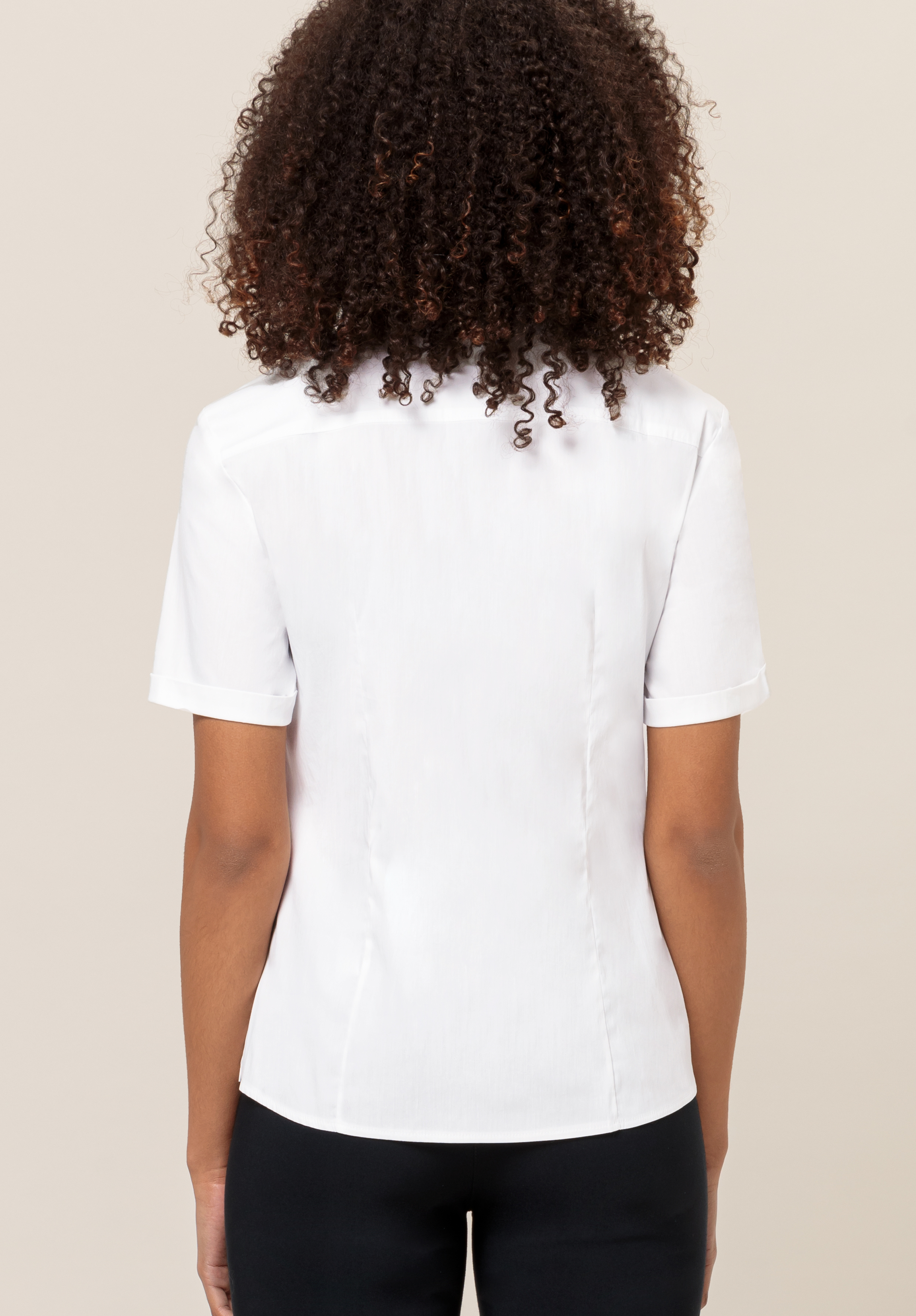 Eine Frau mit lockigem Haar ist von hinten zu sehen; sie trägt eine kurzärmelige, figurbetonte weiße Bluse und eine schwarze Hose und steht vor einem schlichten, hellen Hintergrund.