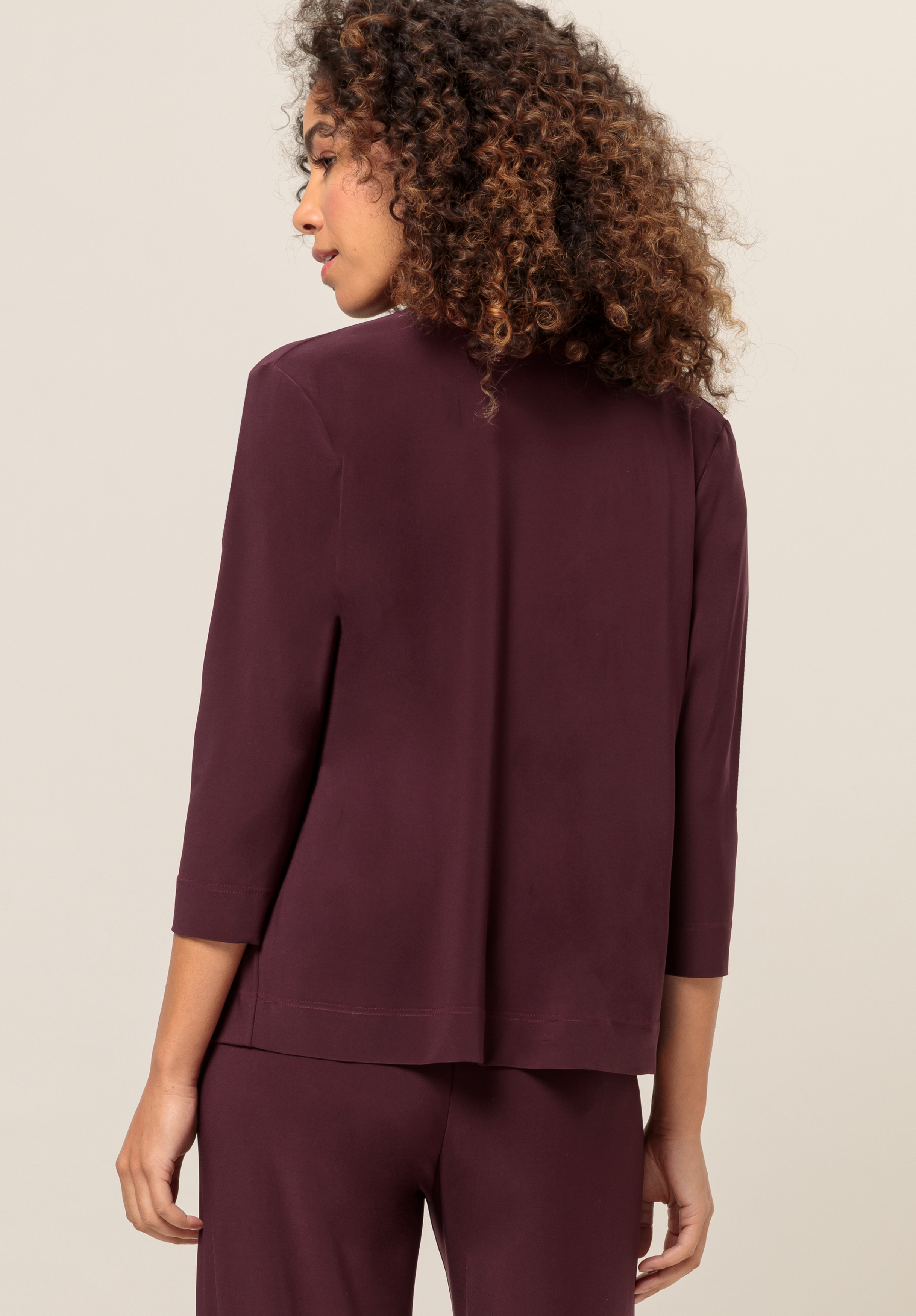 Eine Frau mit lockigem Haar trägt eine dunkelburgunderfarbene Jacke mit Dreiviertelärmeln und eine passende Hose, von hinten vor einem neutralen Hintergrund gezeigt.