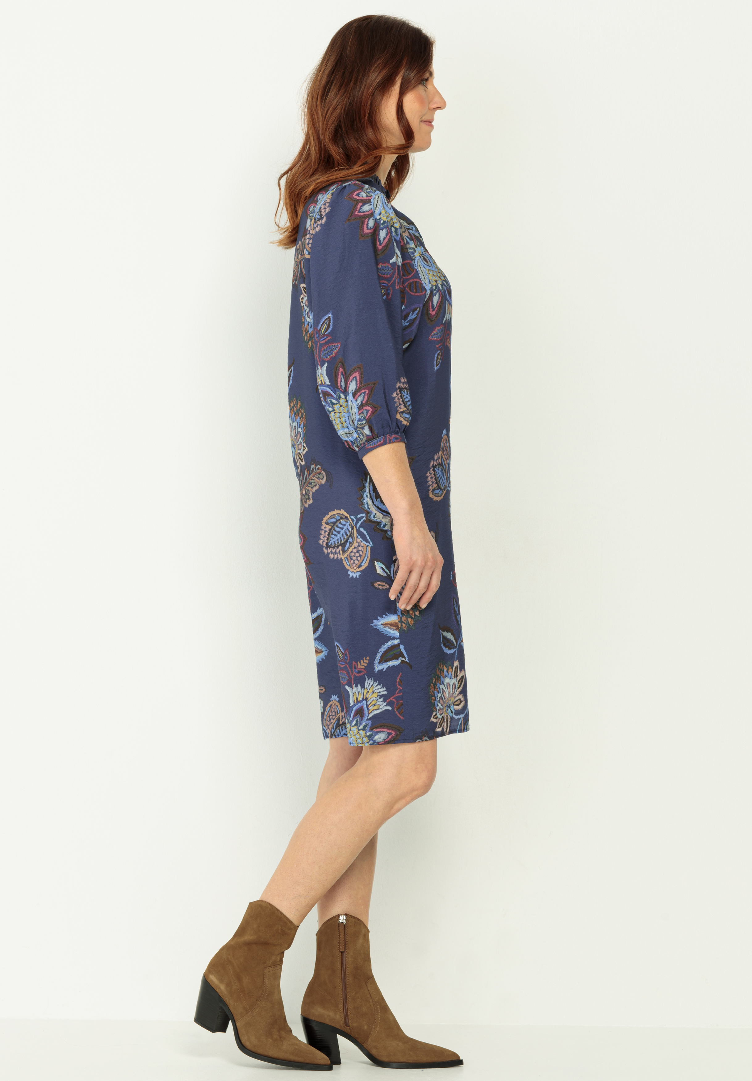 Eine Frau mit gewelltem kastanienbraunem Haar steht seitlich und trägt ein knielanges, blaues Blumenkleid mit Dreiviertelärmeln und braune Stiefeletten vor einem schlichten weißen Hintergrund.