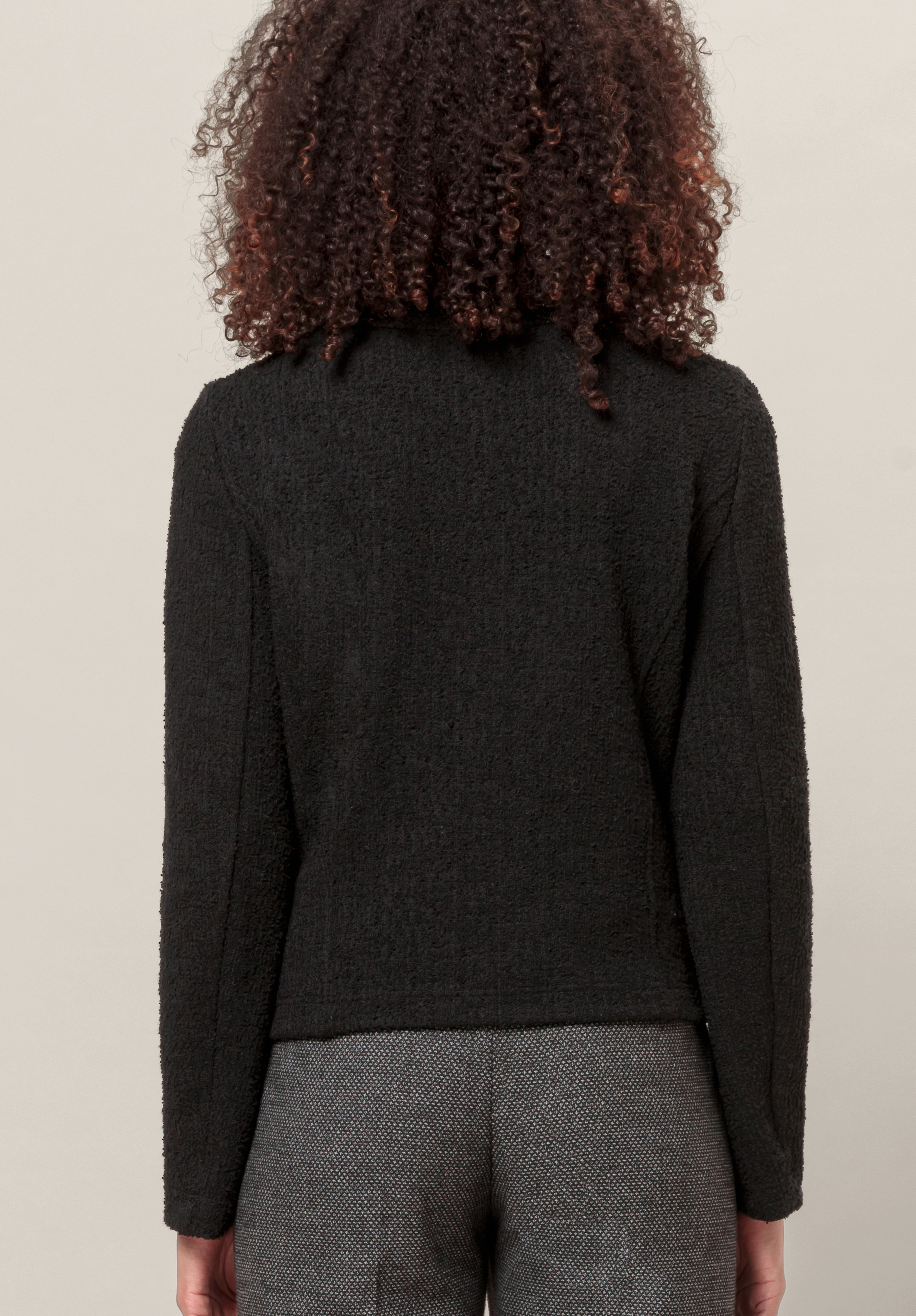 Von hinten ist eine Person mit lockigem Haar zu sehen, die ein strukturiertes schwarzes Langarmoberteil und eine graue Hose trägt und vor einem neutralen Hintergrund steht.