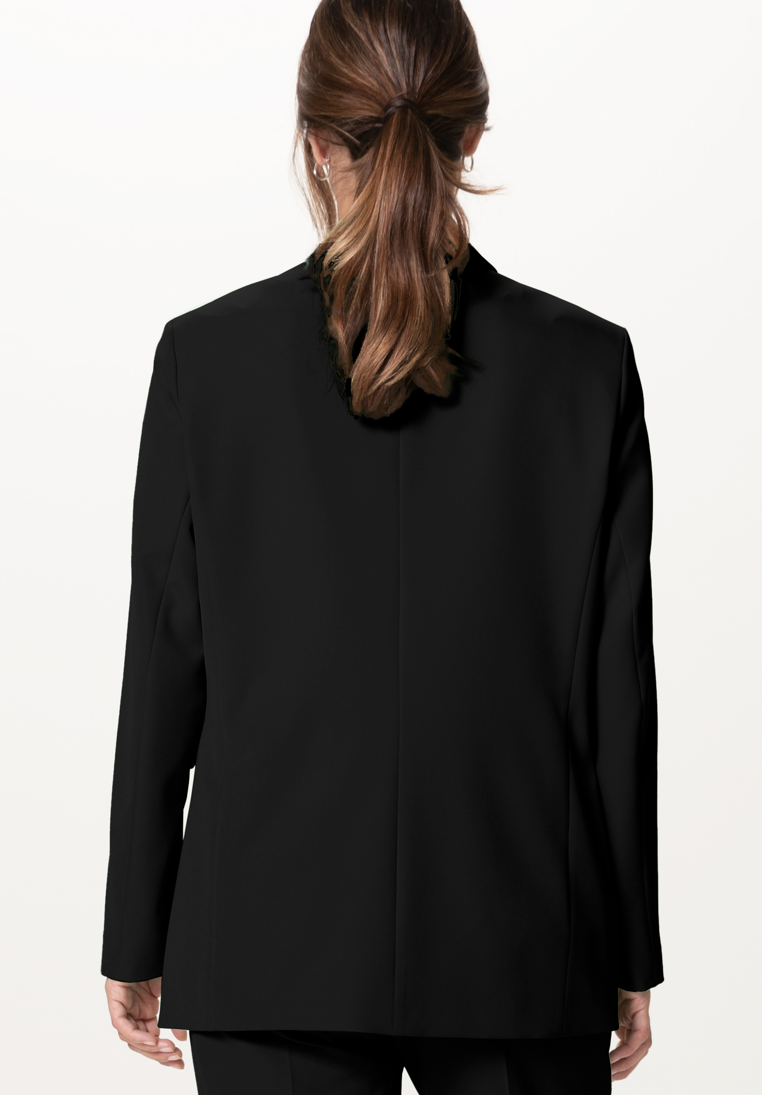 Eine Person mit braunem Haar in einem niedrigen Pferdeschwanz ist von hinten zu sehen. Sie trägt einen schwarzen Blazer und eine schwarze Hose vor einem schlichten hellen Hintergrund.