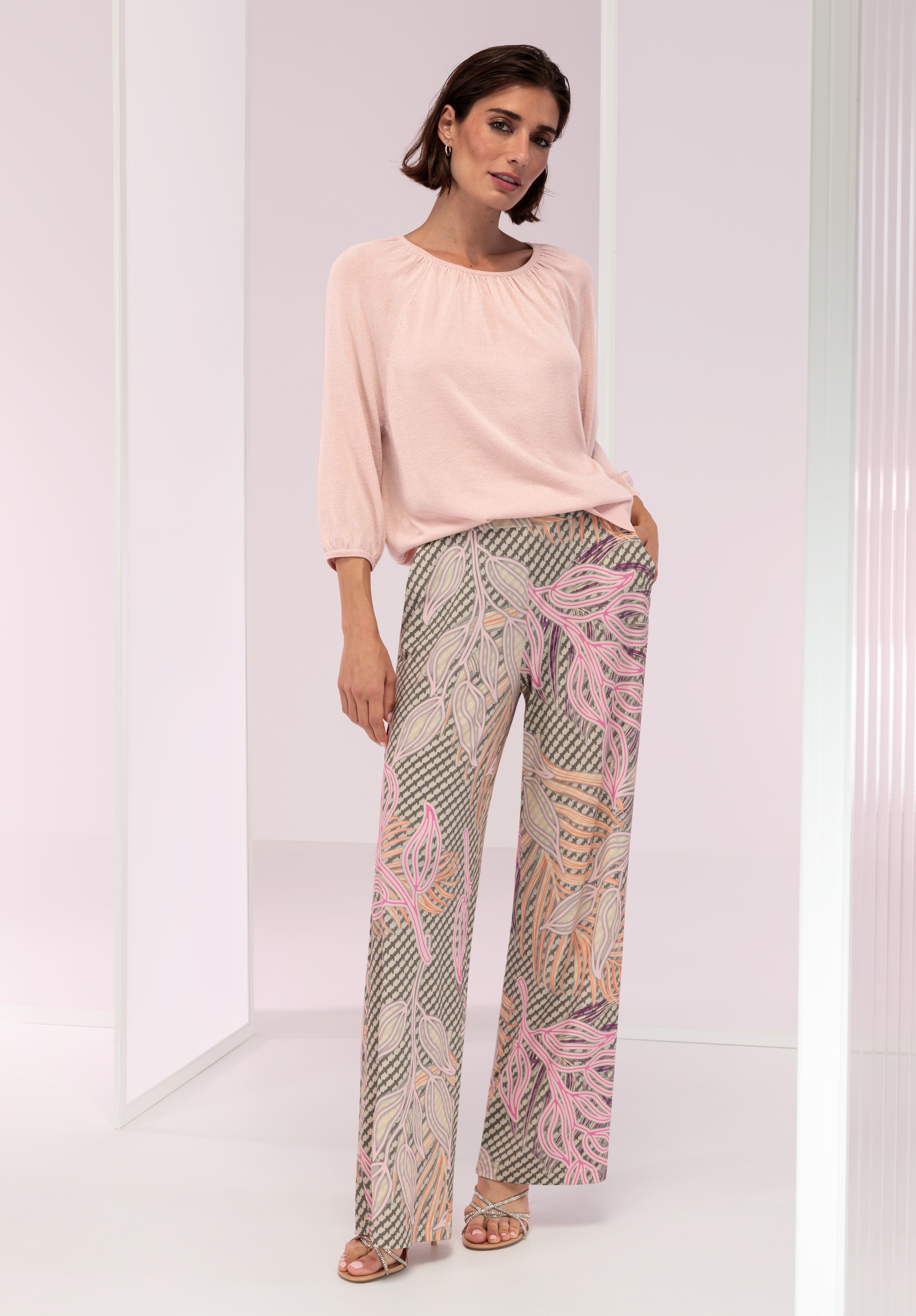 Eine Frau steht in einem hellen, modernen Raum und trägt eine blassrosa Bluse und weite, gemusterte Hosen mit rosa, beigen und grauen Blattmustern. Sie hat kurzes braunes Haar und trägt Sandalen.