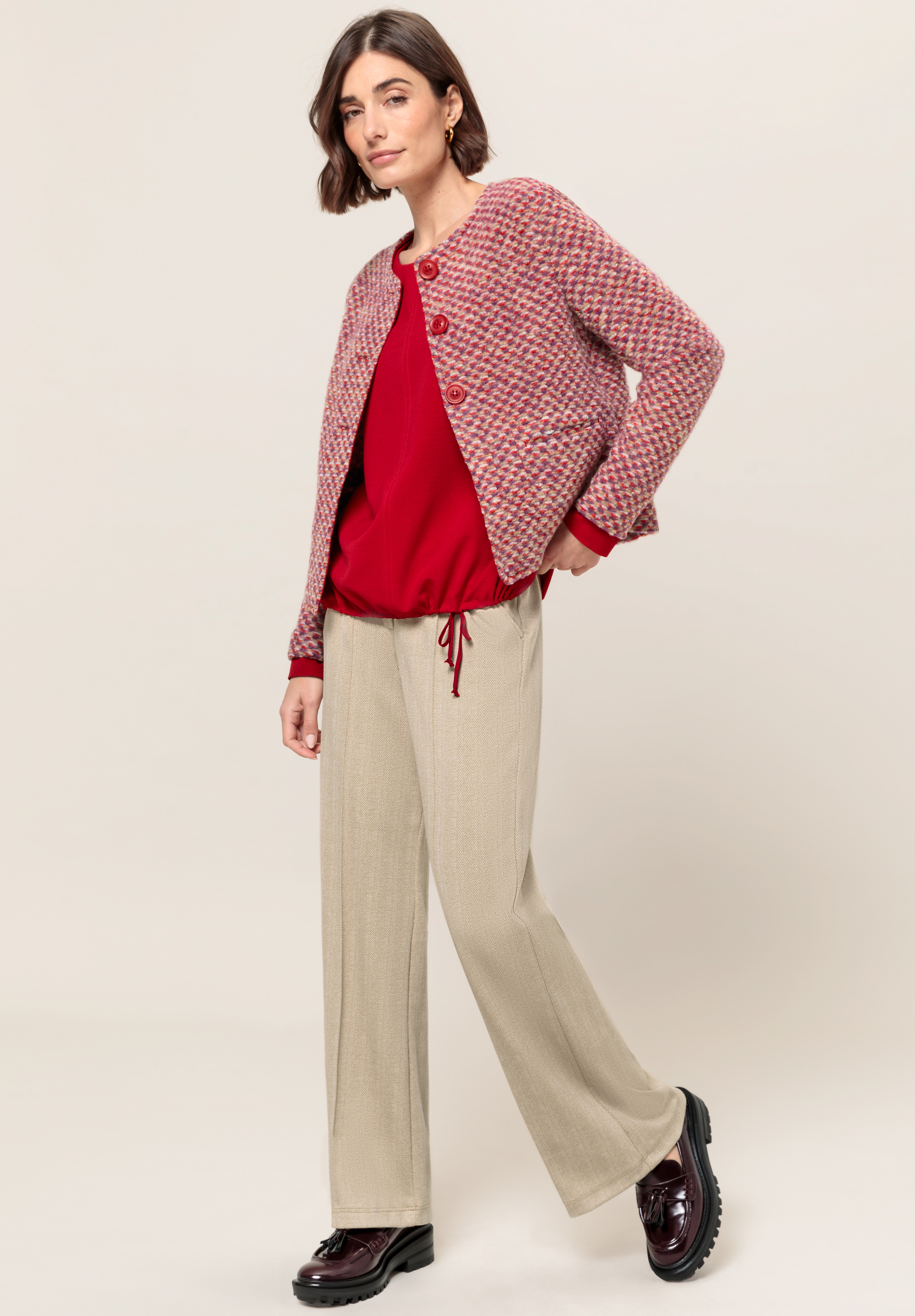 Eine Frau trägt eine rot-pinke Strukturjacke über einem roten Oberteil, dazu beige Hosen mit weitem Bein und dunkelburgunderfarbene Schuhe. Sie steht vor einem schlichten hellen Hintergrund und blickt selbstbewusst in die Kamera.