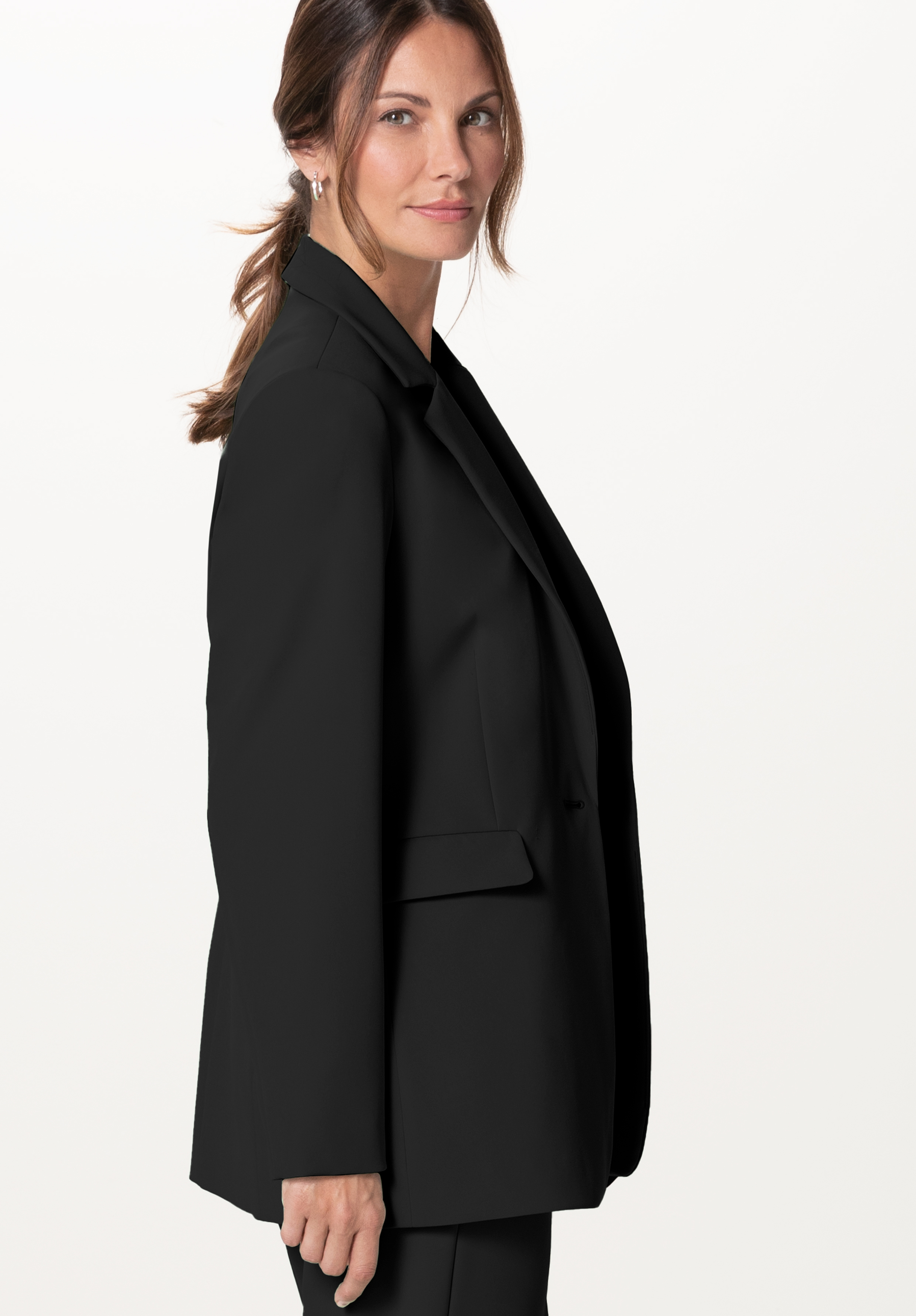 Eine Frau mit braunen Haaren, die zu einem tief sitzenden Pferdeschwanz zusammengebunden sind, trägt einen schwarzen Blazer und eine Hose; sie steht seitlich vor der Kamera und lächelt vor einem schlichten weißen Hintergrund.