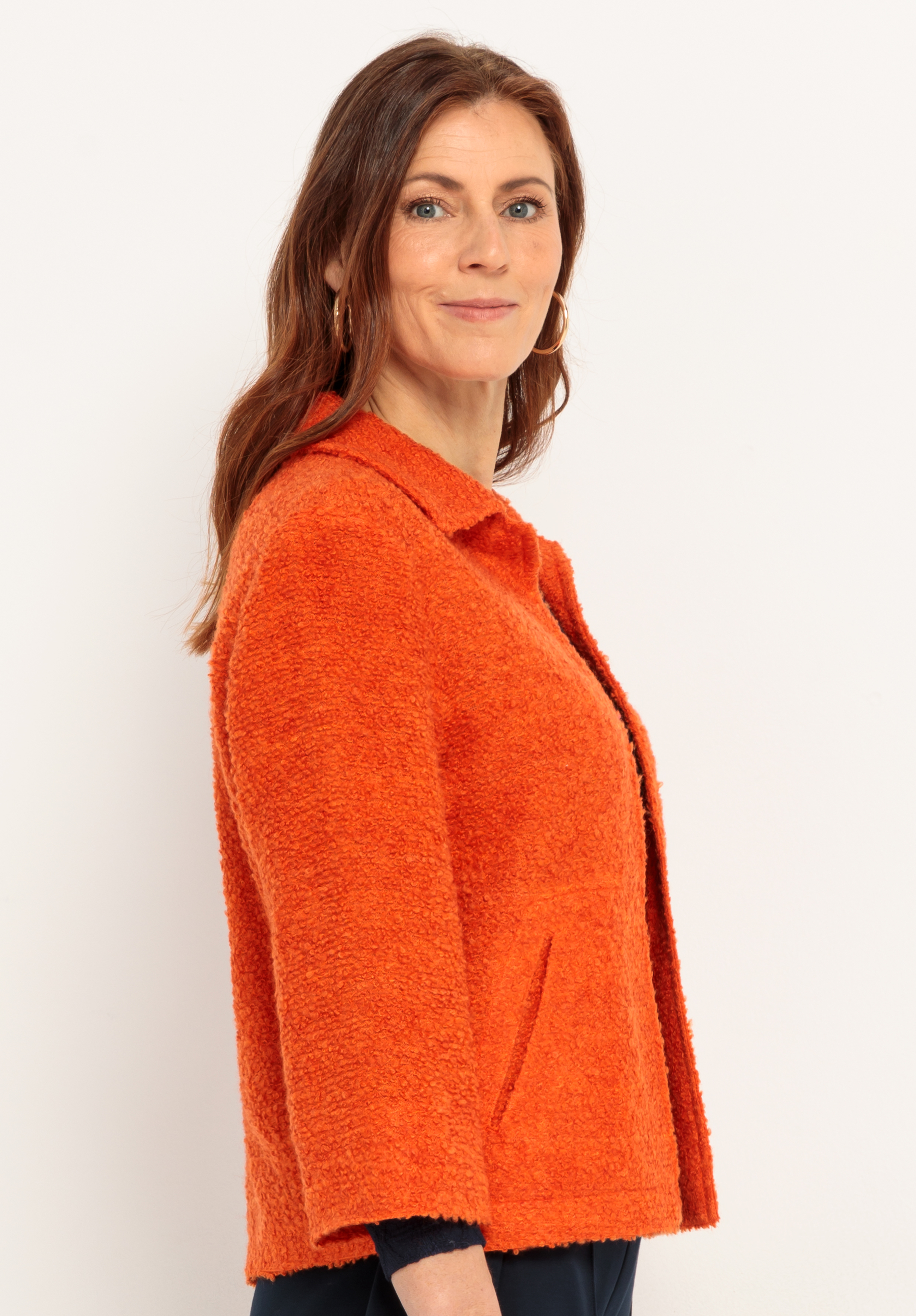 Eine Frau mit langen braunen Haaren, die eine leuchtend orangefarbene strukturierte Jacke und Ohrringe trägt, steht vor einem schlichten weißen Hintergrund und blickt mit einem leichten Lächeln in die Kamera.