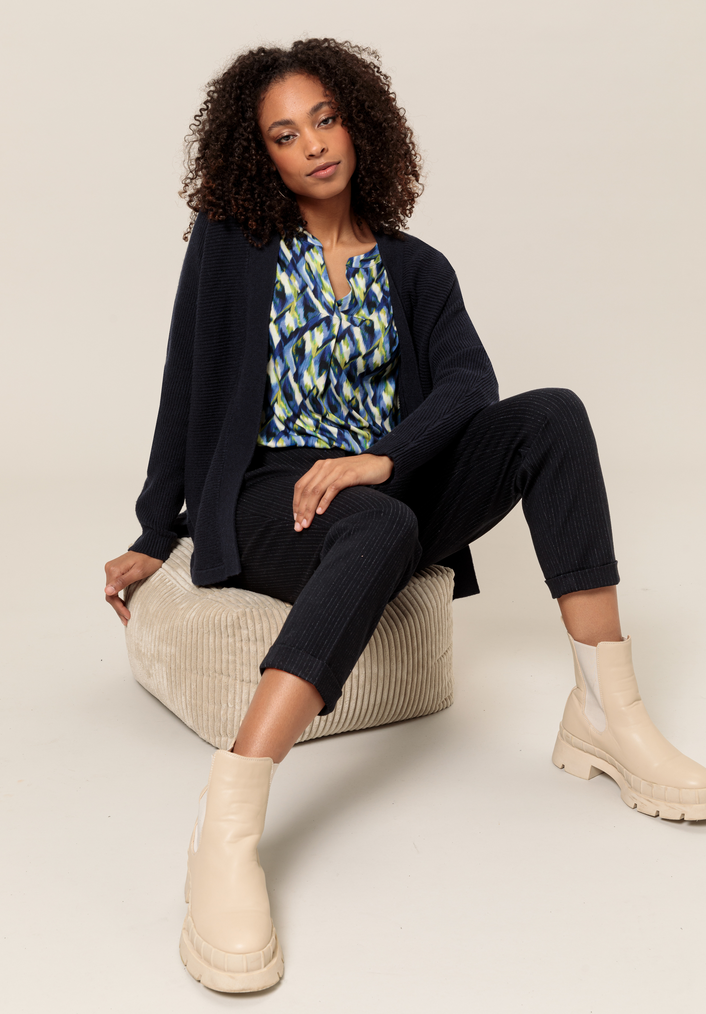 Eine Frau mit lockigem Haar sitzt auf einem beigen Kissen. Sie trägt eine marineblaue Strickjacke, eine gemusterte blaue Bluse, eine schwarze Hose und klobige beige Stiefel vor einem schlichten hellen Hintergrund.