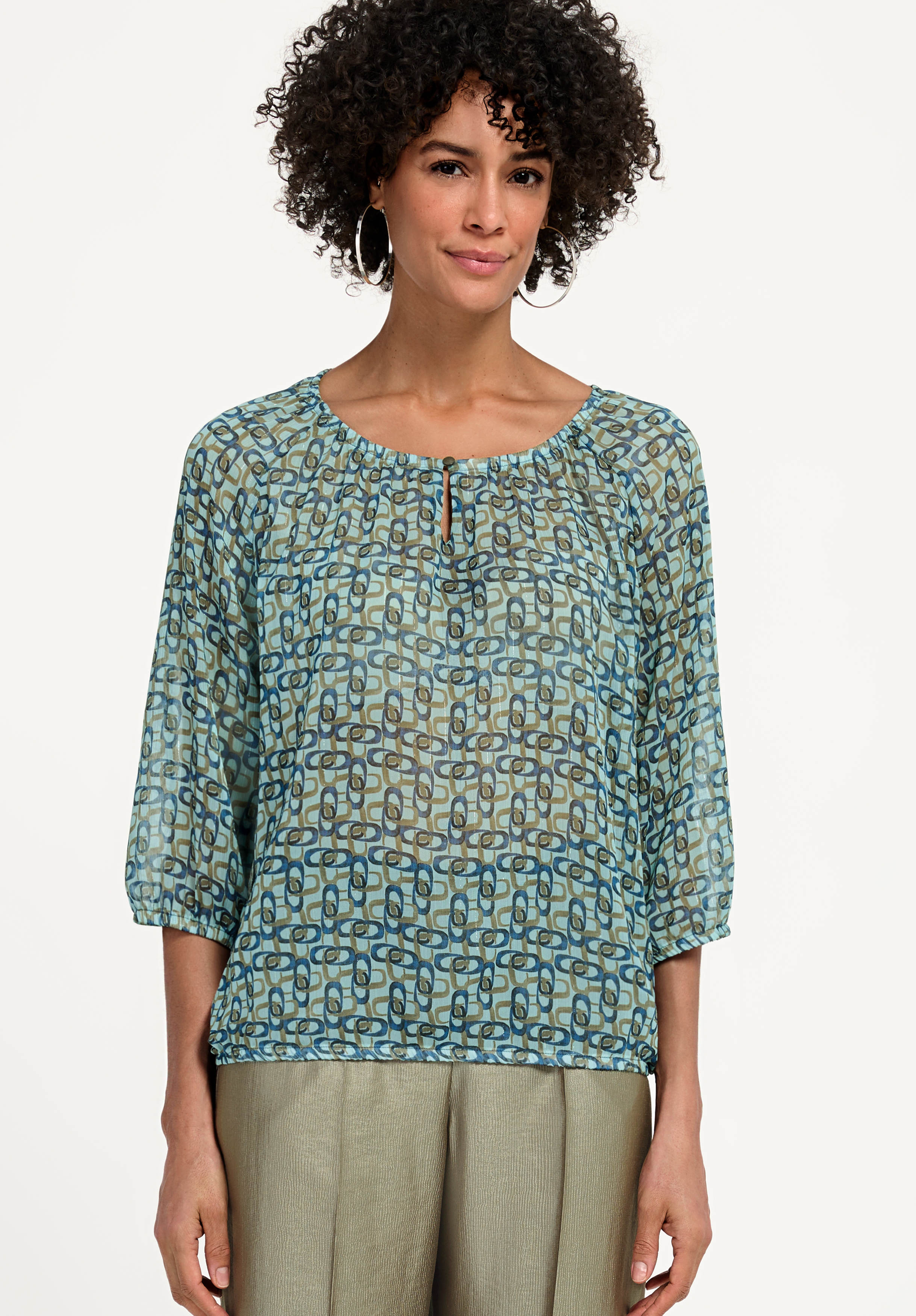 Eine Frau sitzt auf einem weißen Hocker. Sie trägt ein blau gestreiftes Hemd, blaue Jeans und hellblaue Sandalen mit Absatz. Sie hat lockiges Haar, goldene Ohrringe und blickt vor einem schlichten Hintergrund selbstbewusst in die Kamera.