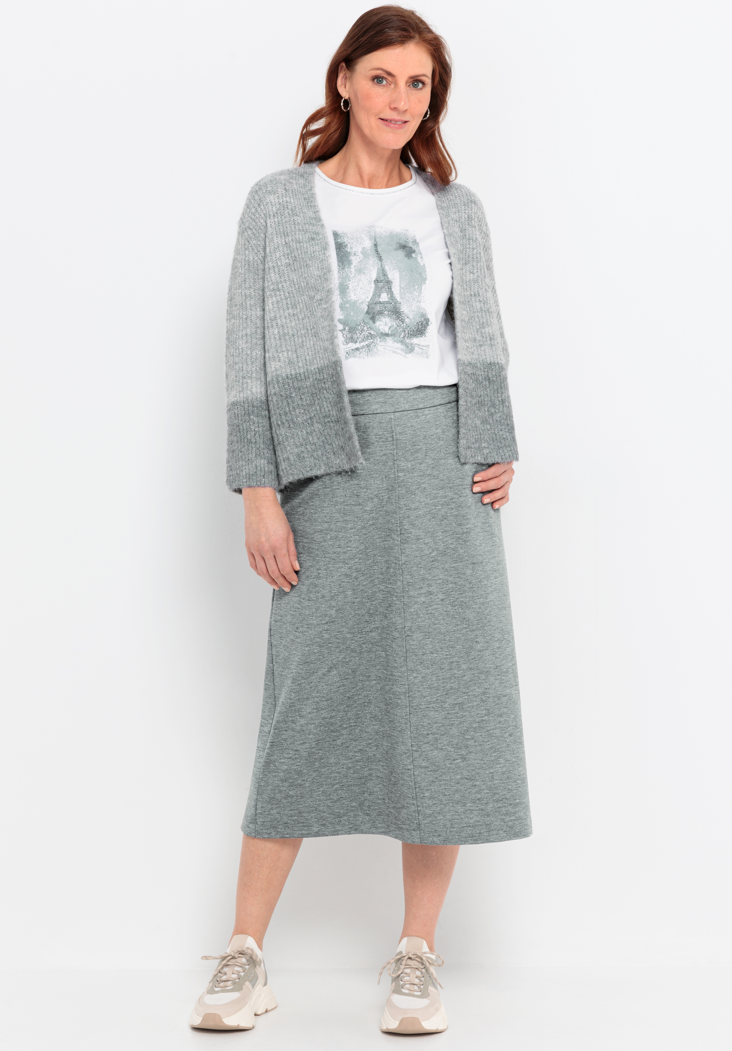 Eine Frau steht vor einem weißen Hintergrund und trägt eine graue Strickjacke, ein weißes T-Shirt mit einer Eiffelturm-Grafik, einen grauen Midi-Rock und beigefarbene Turnschuhe. Sie hat mittellanges braunes Haar und lächelt sanft.