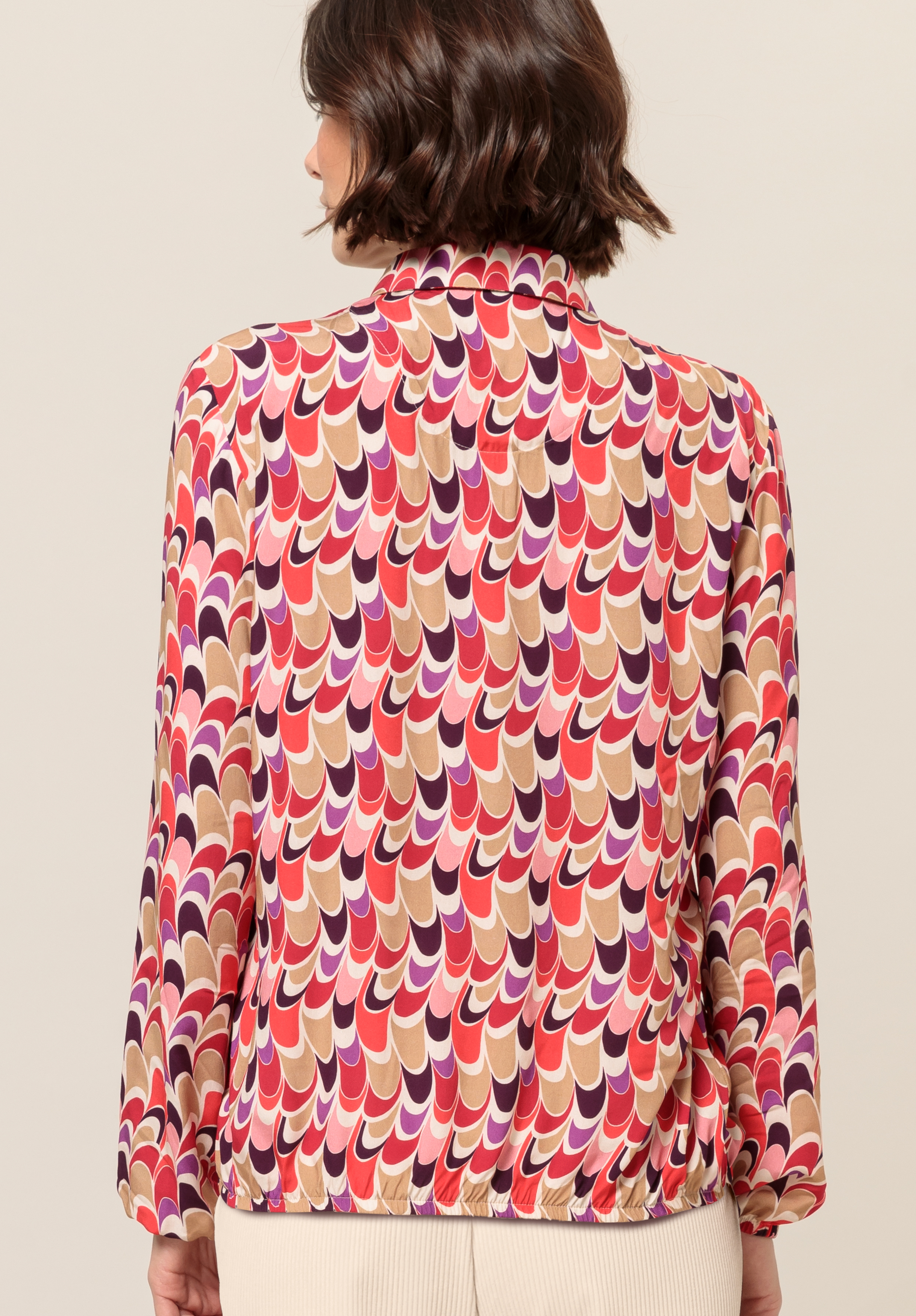 Frau mit kurzen, welligen braunen Haaren, trägt eine langärmelige Bluse mit einem bunten Retro-Wellenmuster in Rot-, Rosa-, Lila- und Beigetönen; von hinten fotografiert.