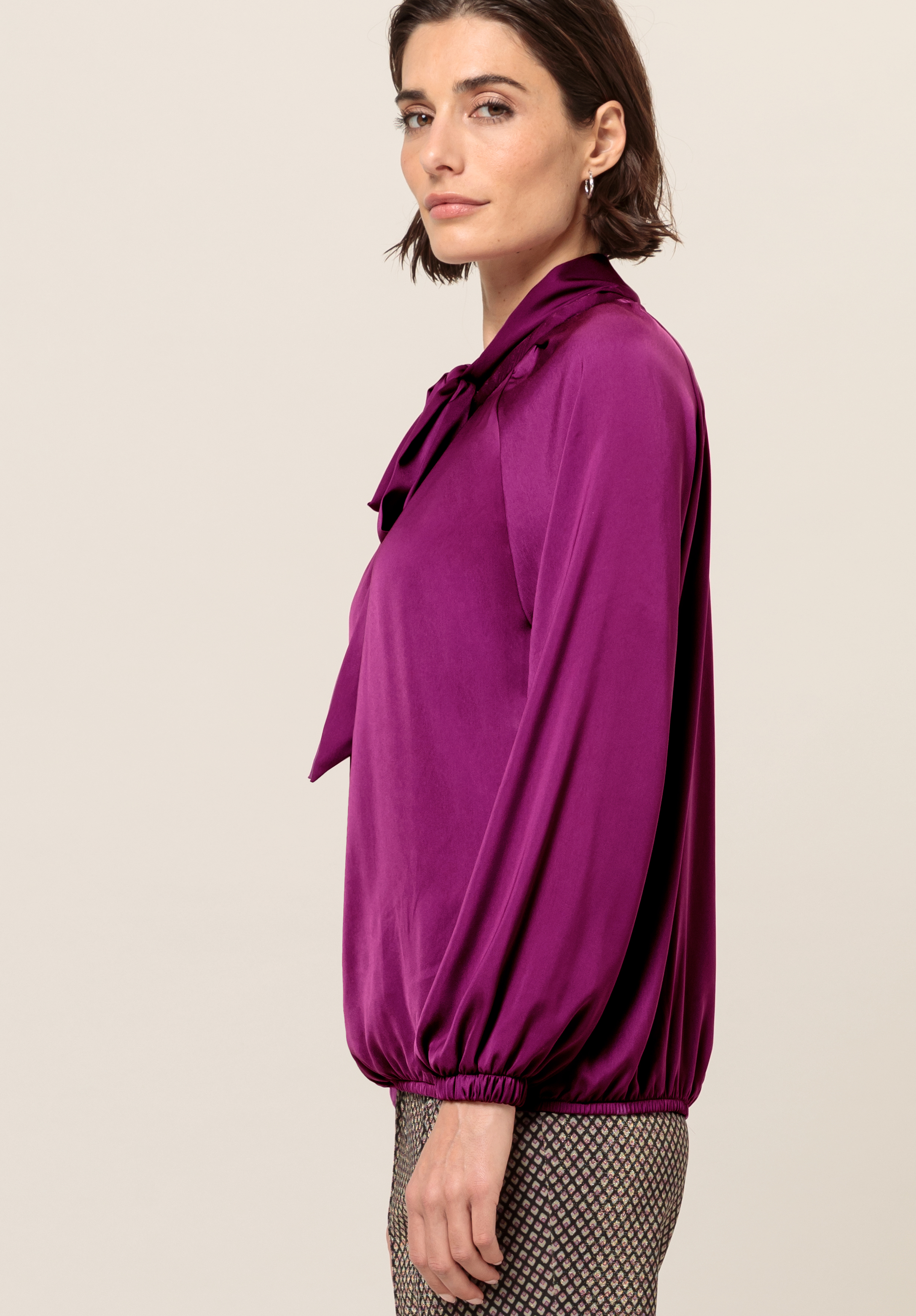 Eine Frau mit kurzen braunen Haaren trägt eine langärmelige, violette Bluse mit einer Schleife am Hals und eine gemusterte Hose und steht im Profil vor einem schlichten hellen Hintergrund.