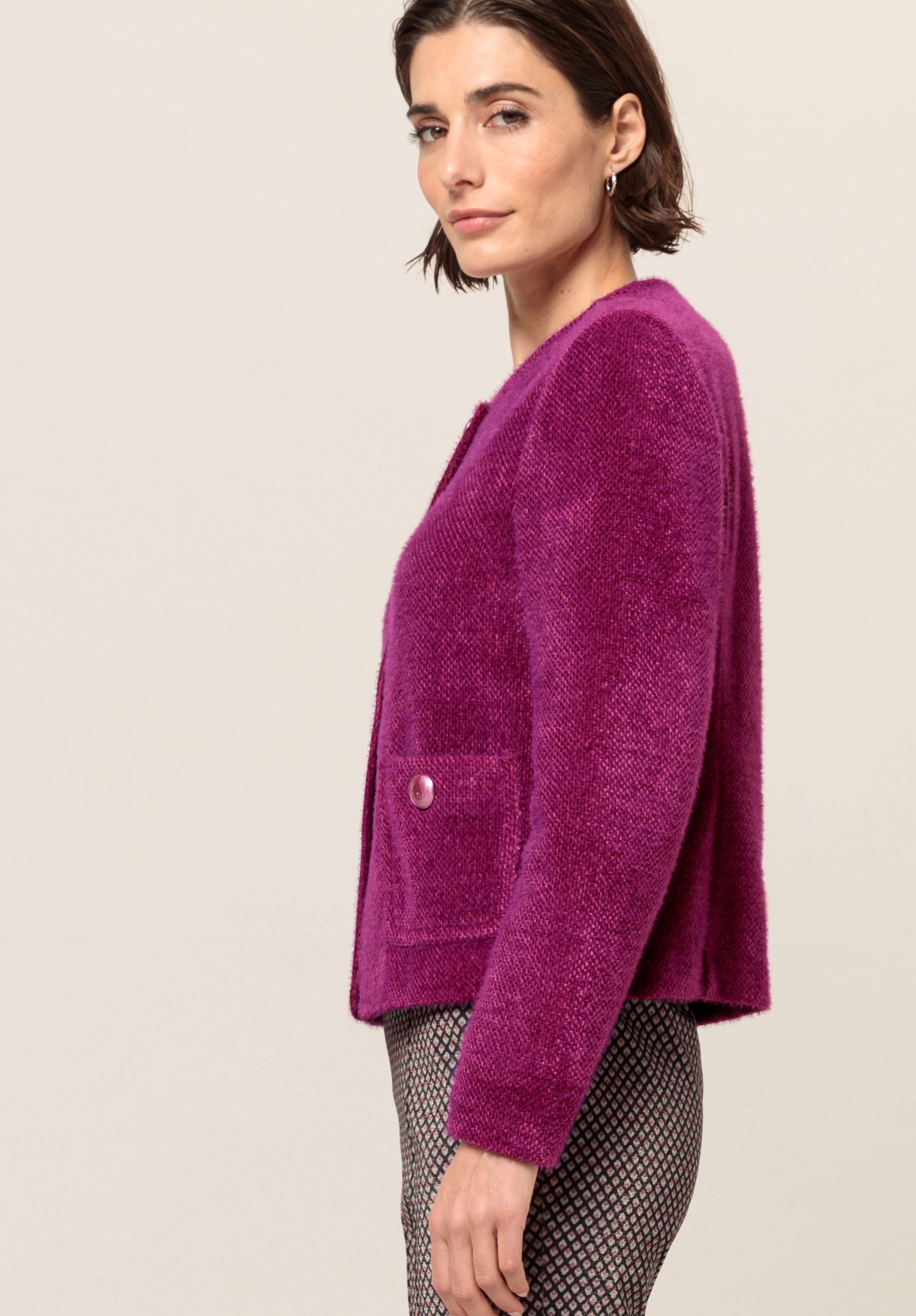 Eine Frau mit kurzen braunen Haaren trägt eine strukturierte, leuchtend violette Jacke mit Vordertaschen und einem Knopf, kombiniert mit gemusterten schwarz-weißen Hosen, und steht seitlich vor einem schlichten Hintergrund.