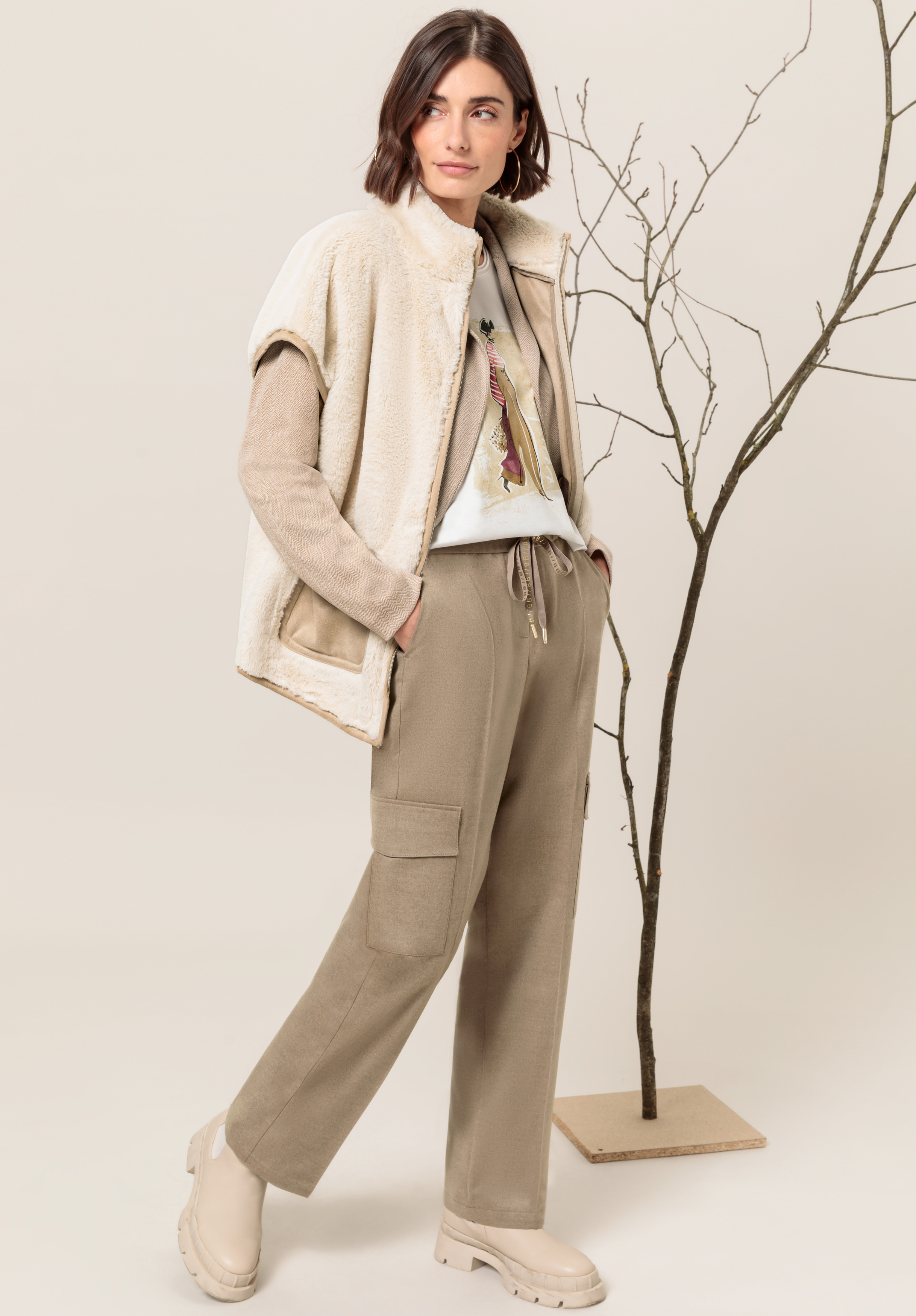 Eine Frau steht vor einem kahlen Dekobaum. Sie trägt beige weite Hosen, ein Sweatshirt mit Grafik, eine cremefarbene Sherpa-Weste und beige Stiefel. Sie hat kurze braune Haare und eine entspannte, selbstbewusste Pose.