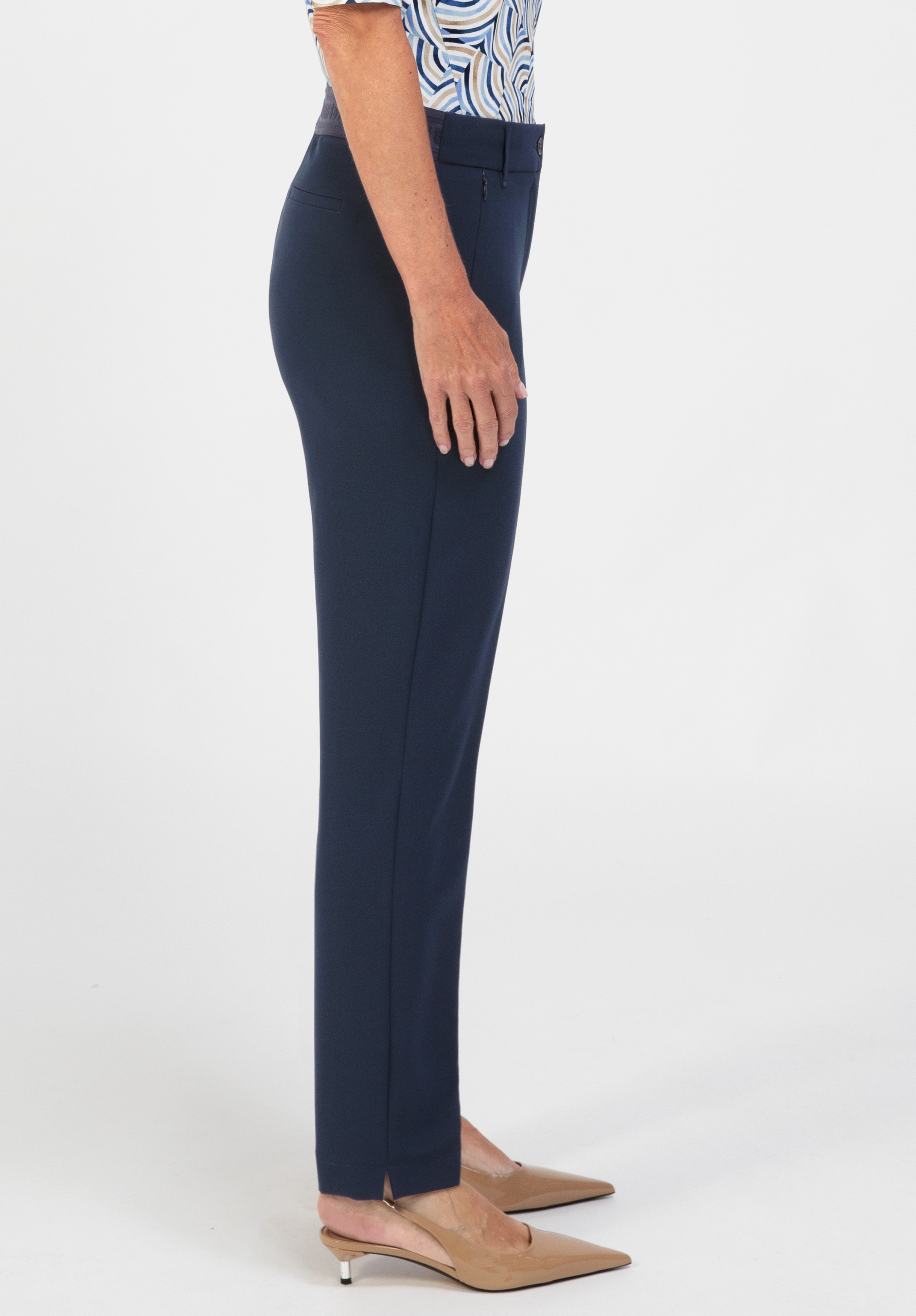 Eine Person, die seitlich steht und eine marineblaue Hose mit geradem Bein, ein gemustertes, in die Hose gestecktes Hemd sowie beige Pumps mit spitzer Zehenpartie trägt, vor einem weißen Hintergrund.