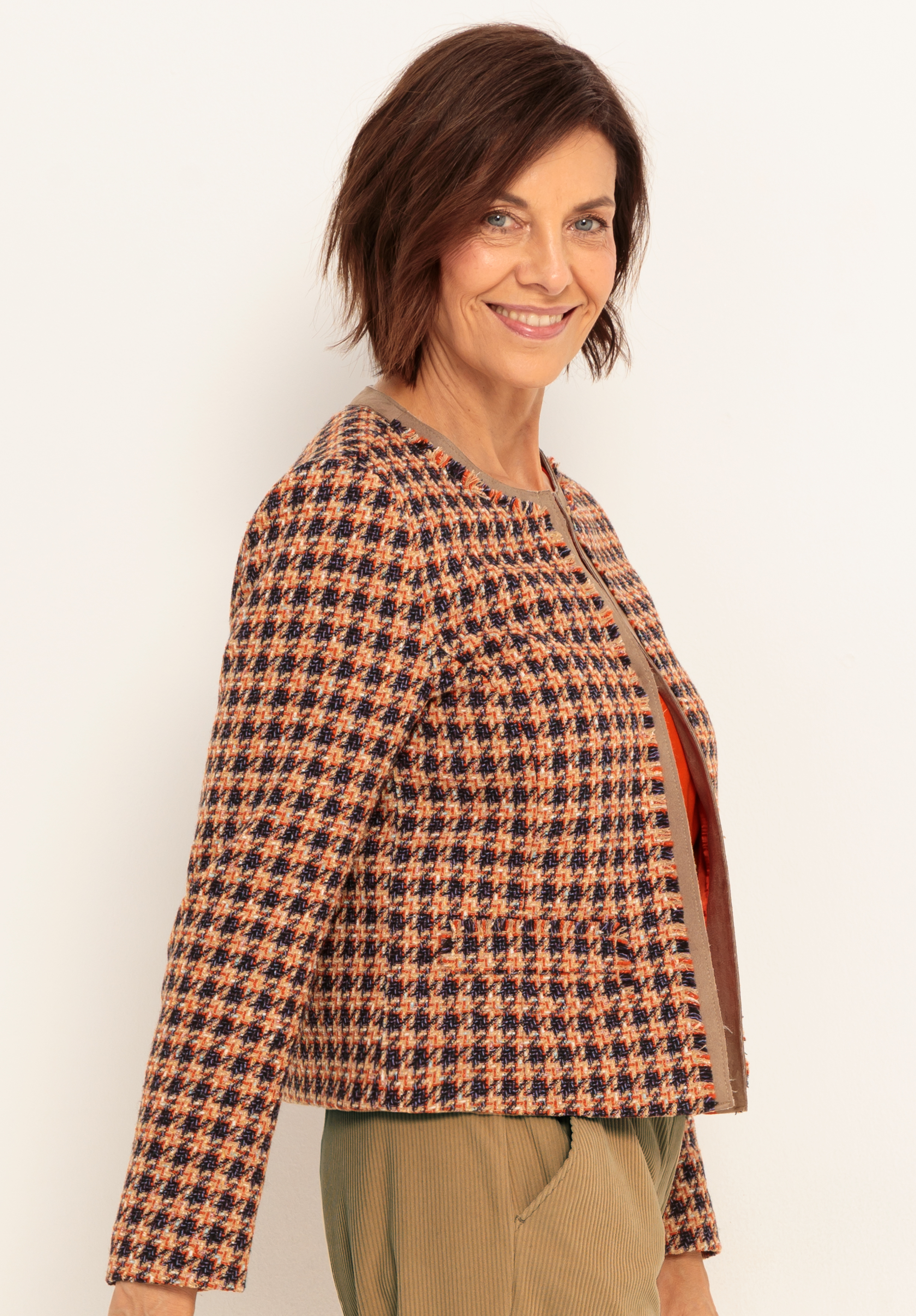 Eine Frau mit kurzen braunen Haaren lächelt, während sie eine gemusterte, karierte Jacke in Braun-, Orange- und Marinetönen über einem rostfarbenen Oberteil und einer hellen Hose vor einem schlichten hellen Hintergrund trägt.